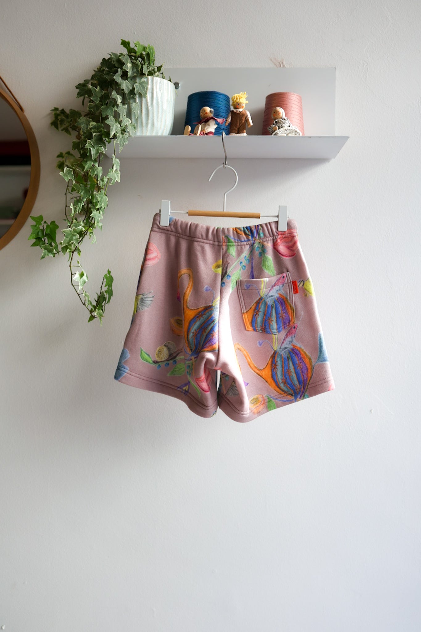 Colorful kids shorts hanging on a white wall with a shelf and plants in the background.