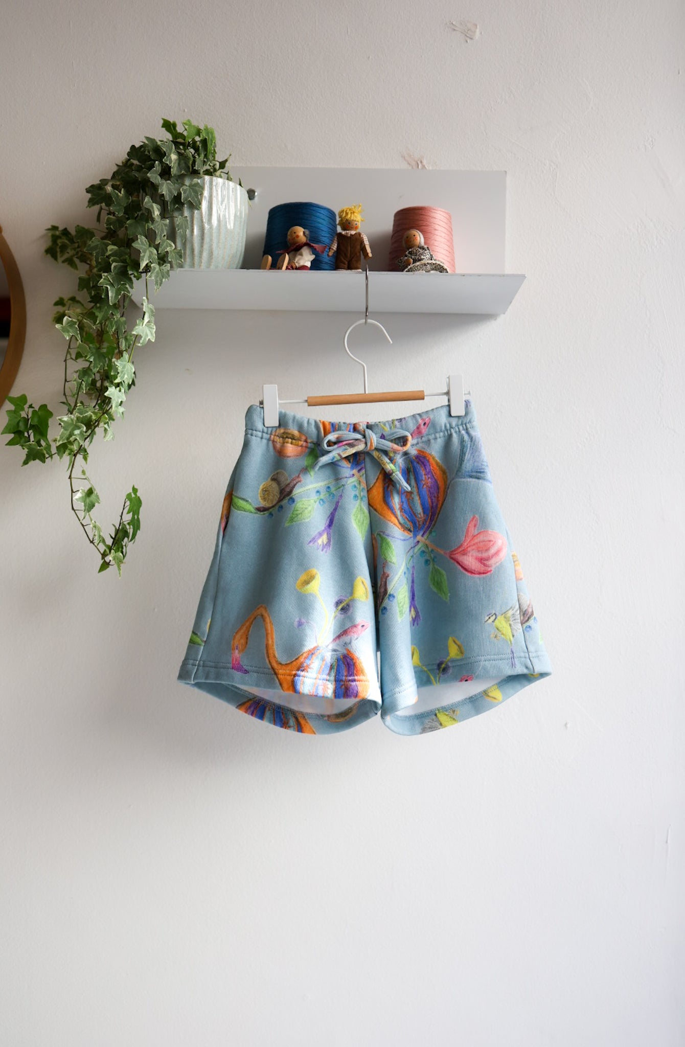 Colorful shorts with floral design hanging on a white wall with a shelf and plant in the background.