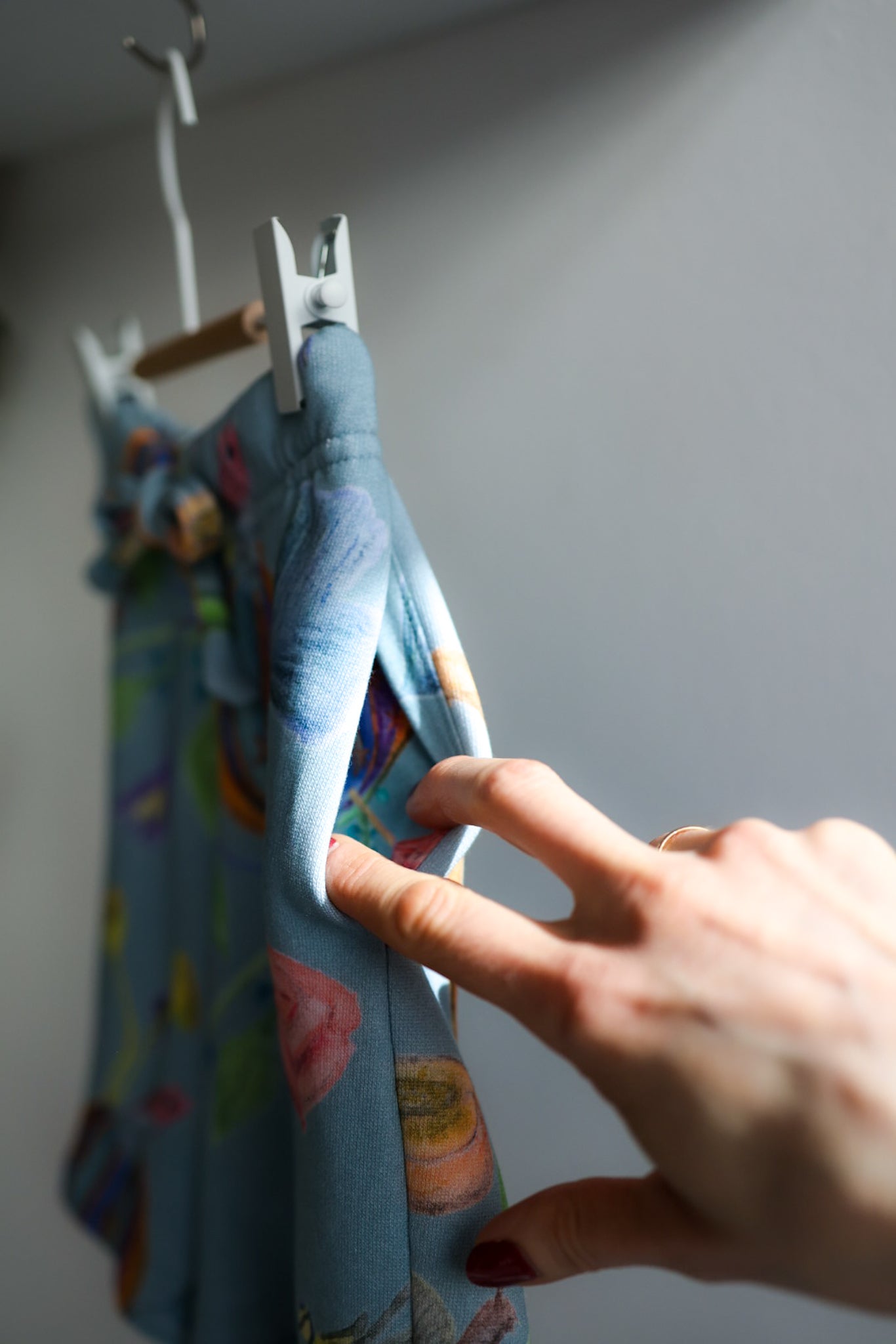 Hand adjusting a blue shorts on a hanger against a gray wall.