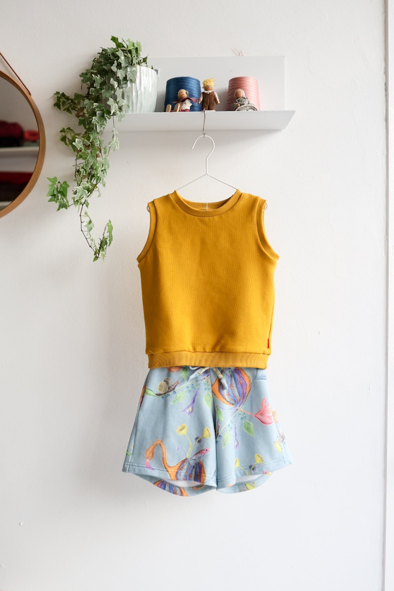 Mustard yellow sleeveless top and colorful shorts on a white wall with decorative elements.