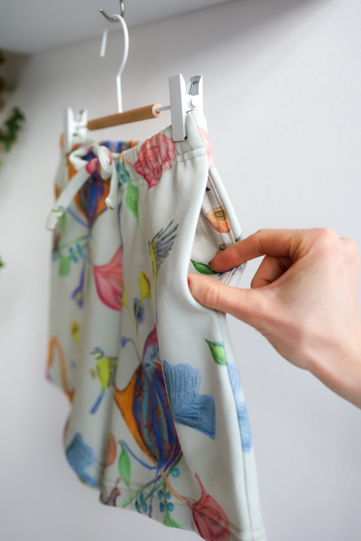 Colorful kids shorts on a hanger with a hand holding it against a white wall.