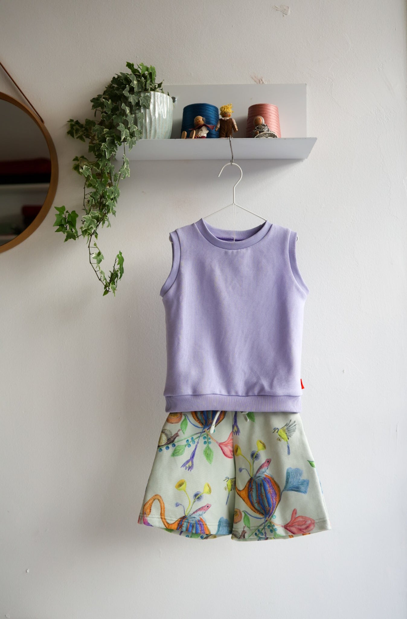Children's outfit with lavender tank top and colorful shorts hanging on a white wall.