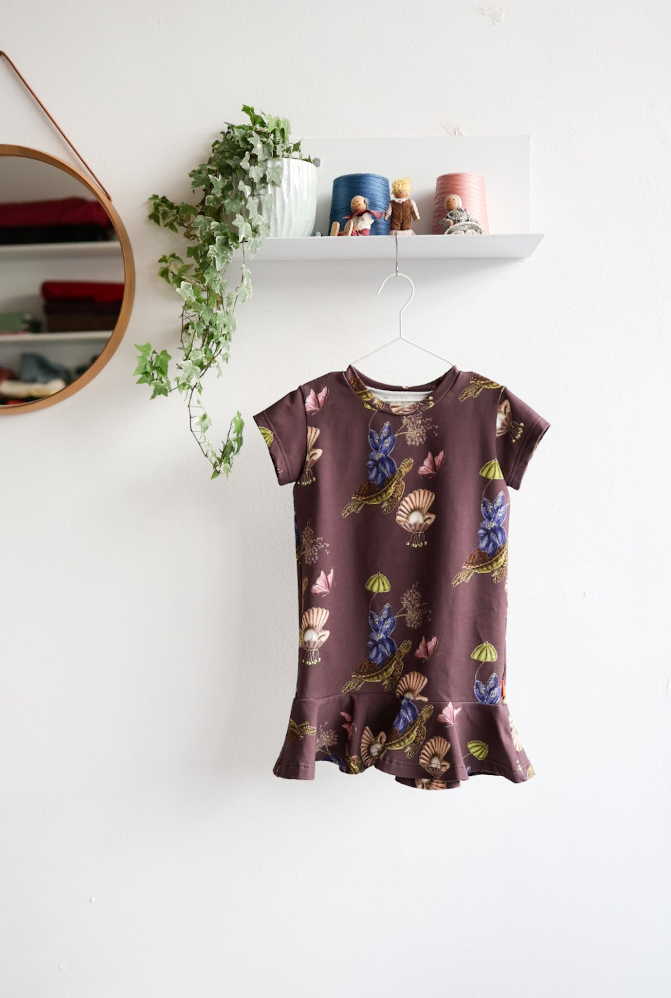 Brown kids dress with nature-themed pattern on a hanger against a white wall.
