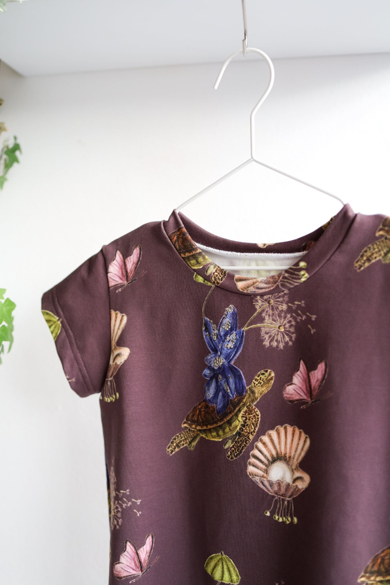 Brown kids dress with nature-themed pattern on a hanger against a white wall.