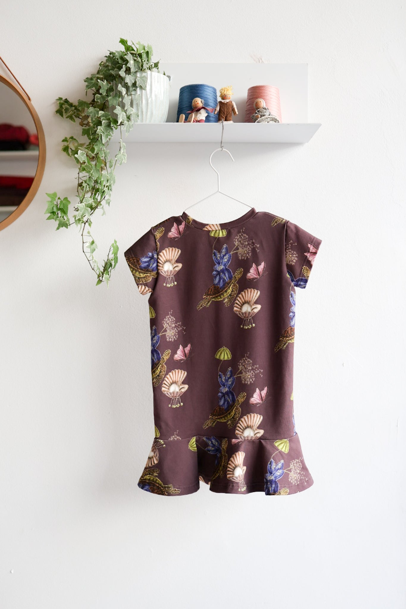 Brown kids dress with nature-themed pattern on a hanger against a white wall.