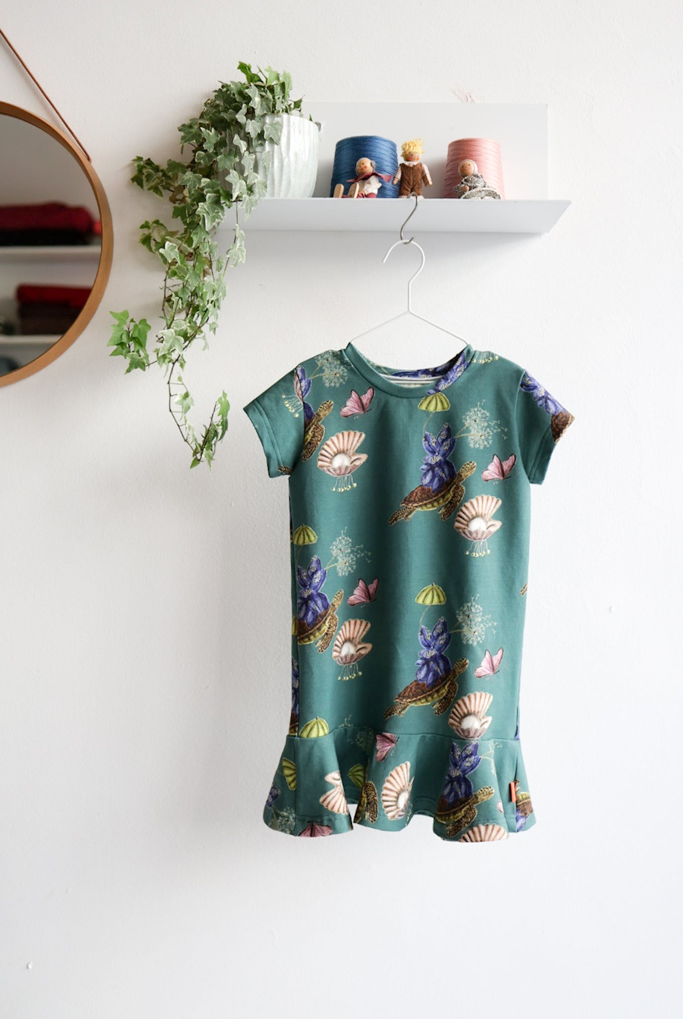 Teal kids dress with nature-themed pattern on a hanger against a white wall.
