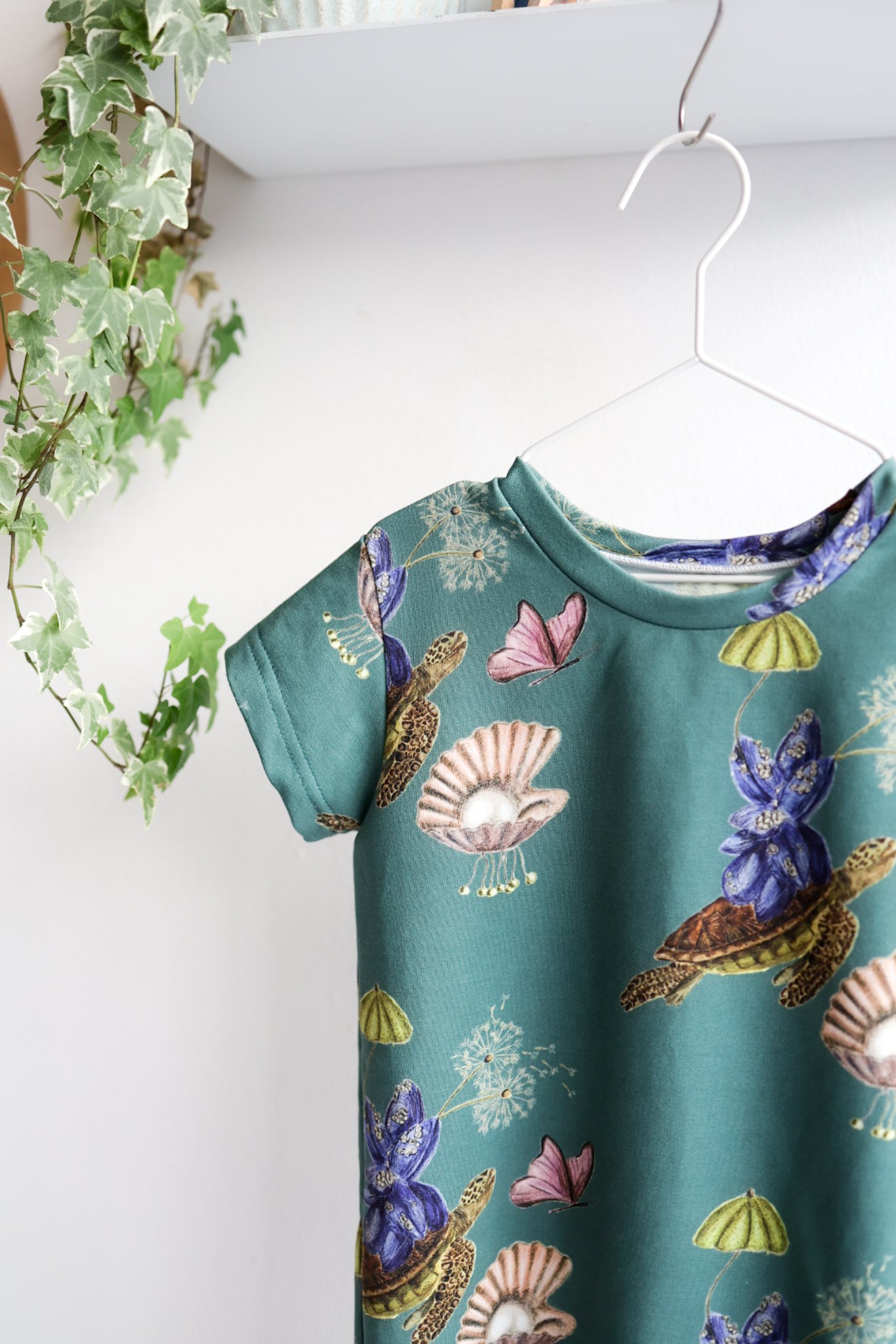 Teal kids dress with nature-themed pattern on a hanger against a white wall.