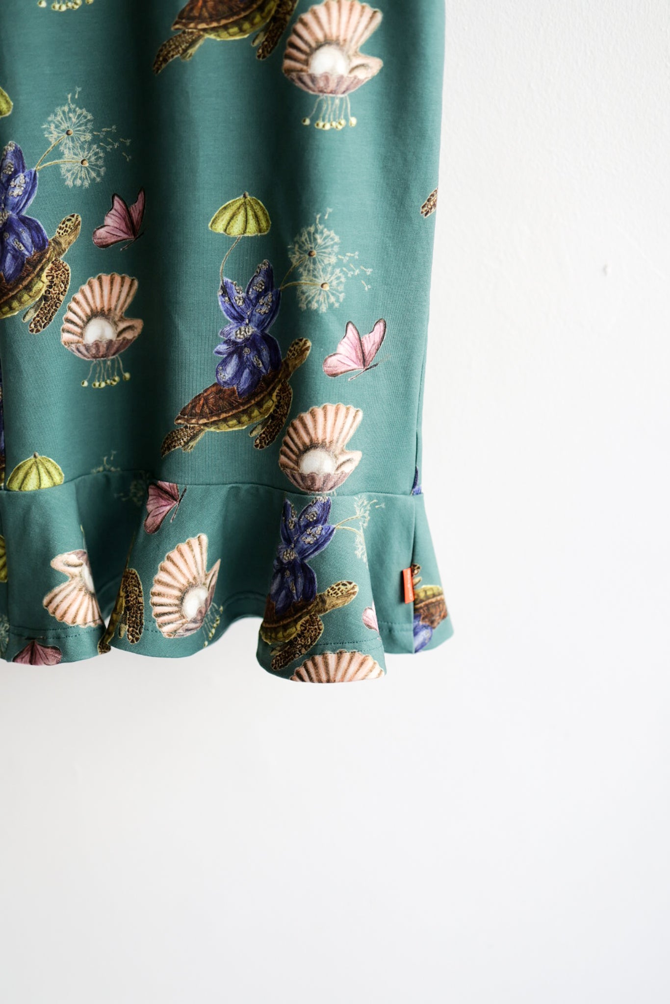 Close-up of a teal kids dress with nature-themed pattern on a hanger against a white wall.
