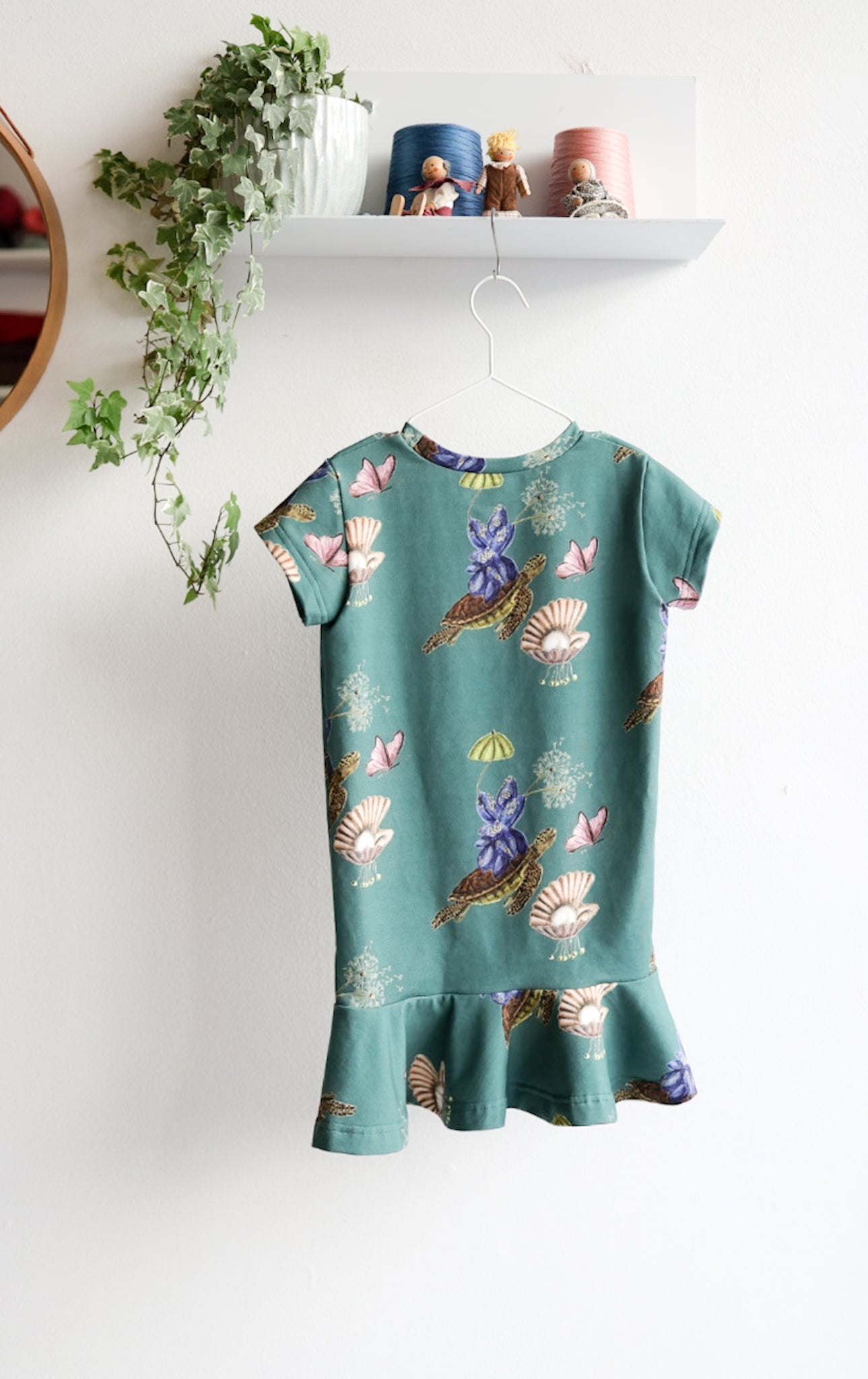 Teal kids dress with nature-themed pattern on a hanger against a white wall.