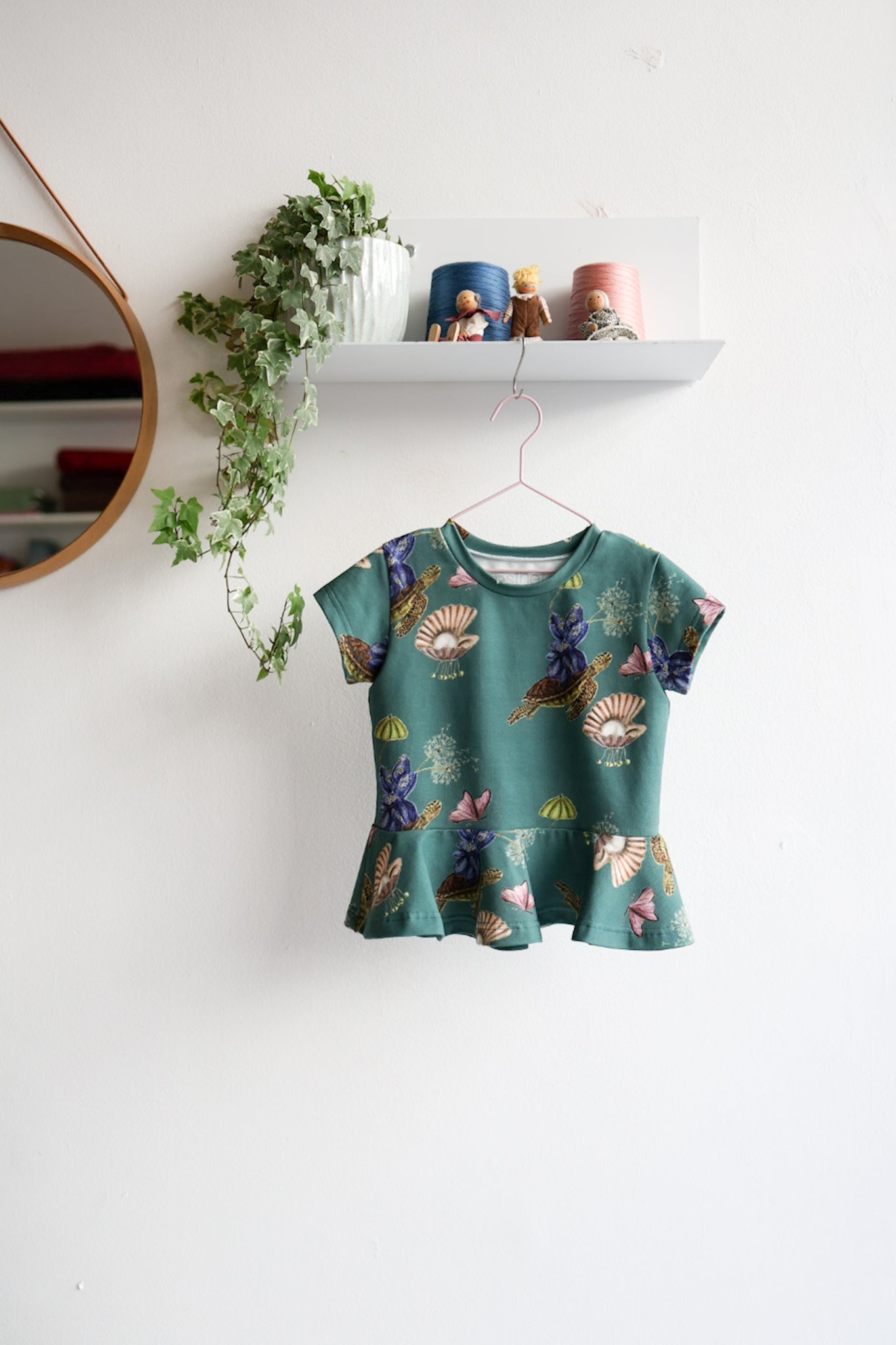 Teal kids top with colorful shell pattern hanging on a white wall.