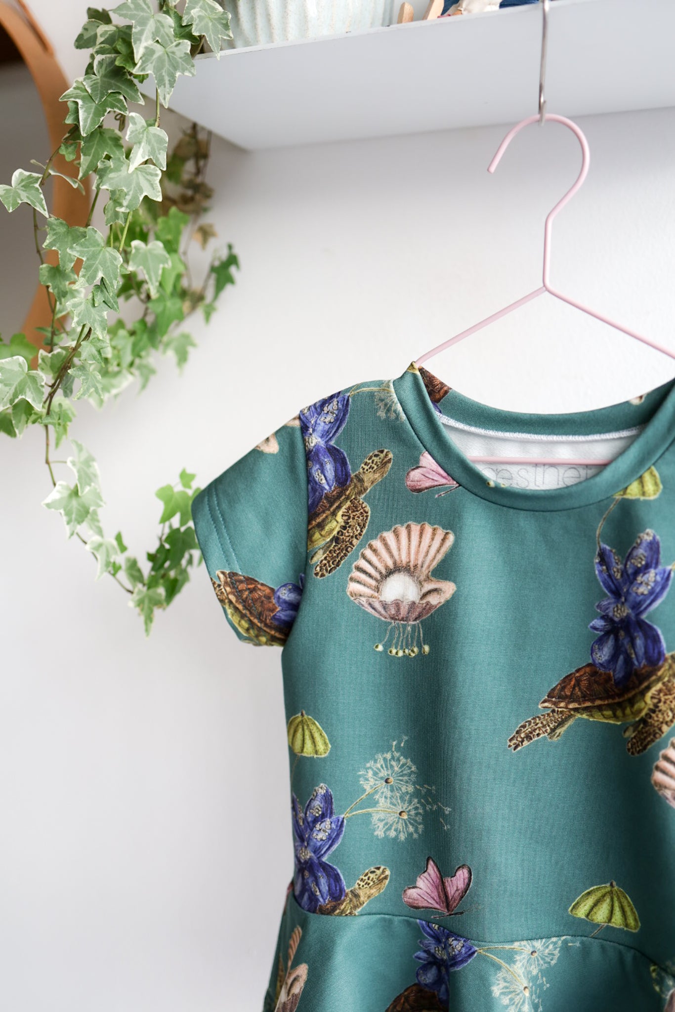 Teal kids top with colorful shell pattern hanging on a white wall.