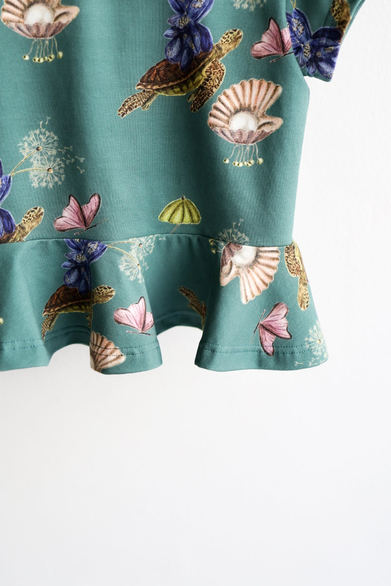Close-up of a teal kids top with colorful shell pattern hanging on a white wall.