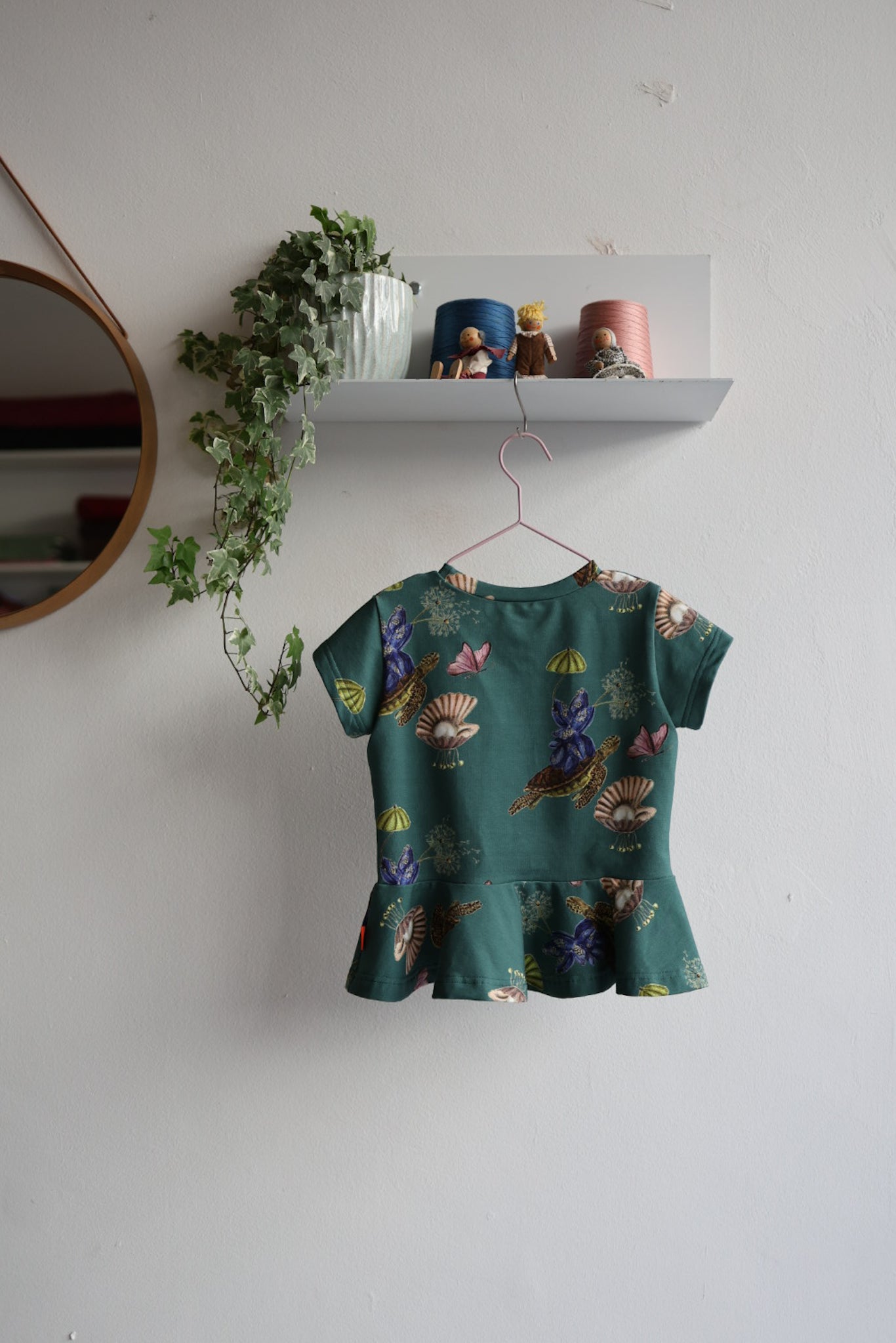Kids nature-themed print teal top hanging on a white wall with decorative items.