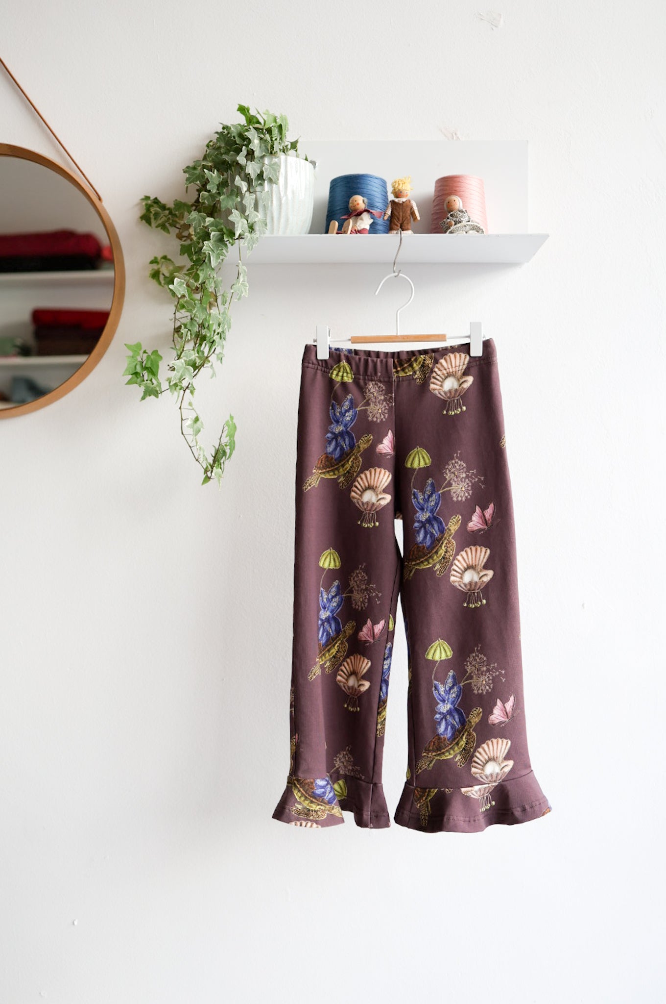 Brown nature-patterned kids pants hanging against a white wall with decorative items.