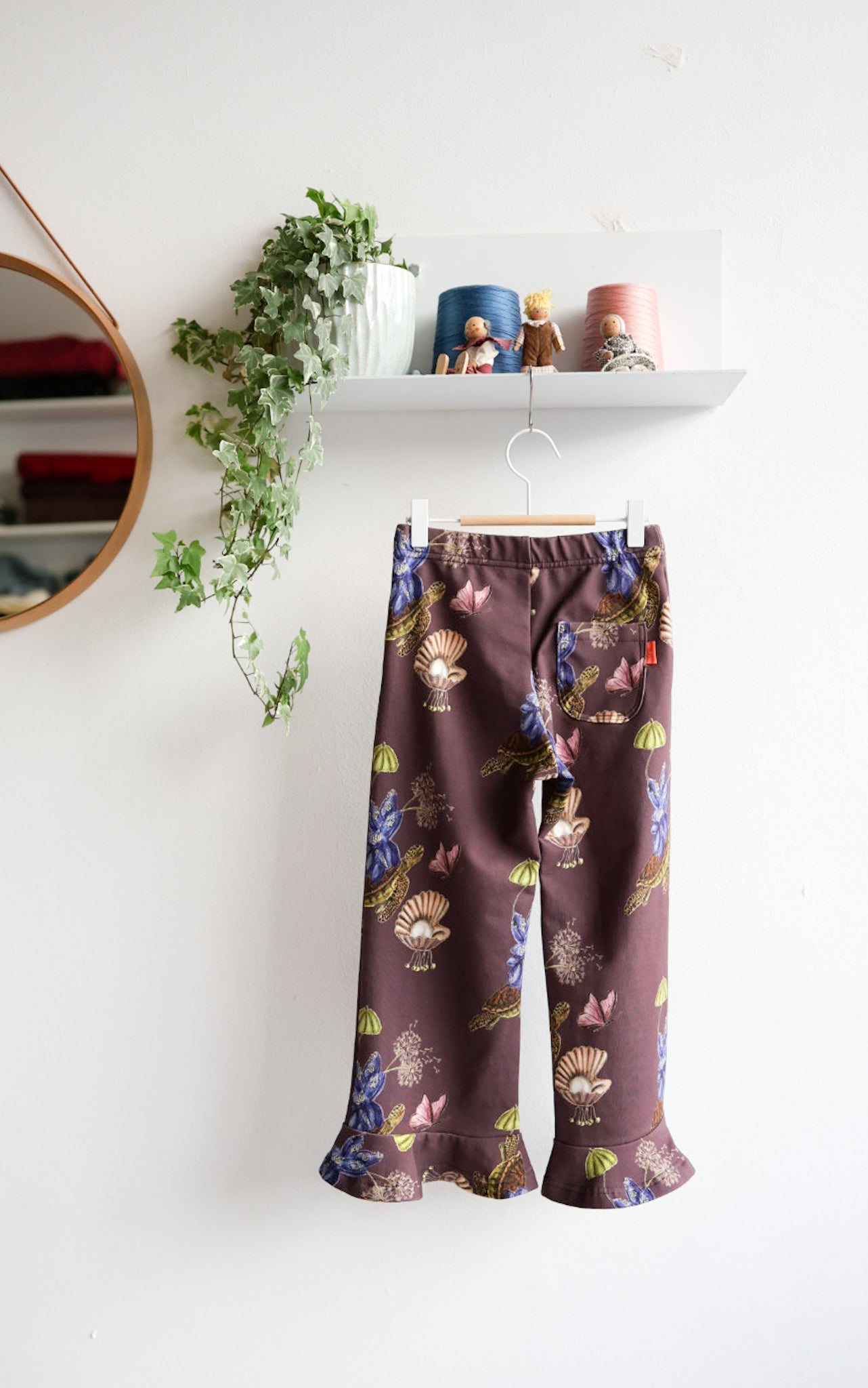 Brown nature-patterned kids pants hanging against a white wall with decorative items.