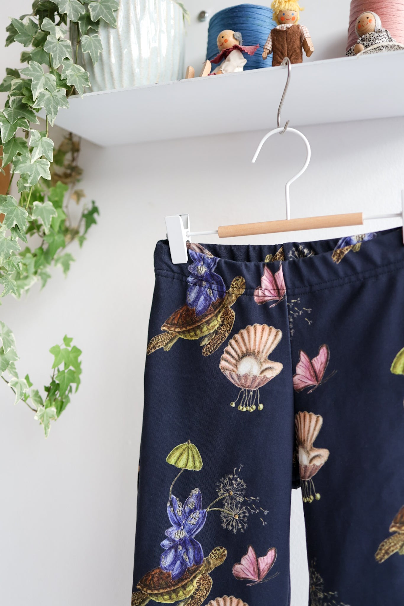 Navy blue nature-patterned kids pants hanging against a white wall with decorative items.