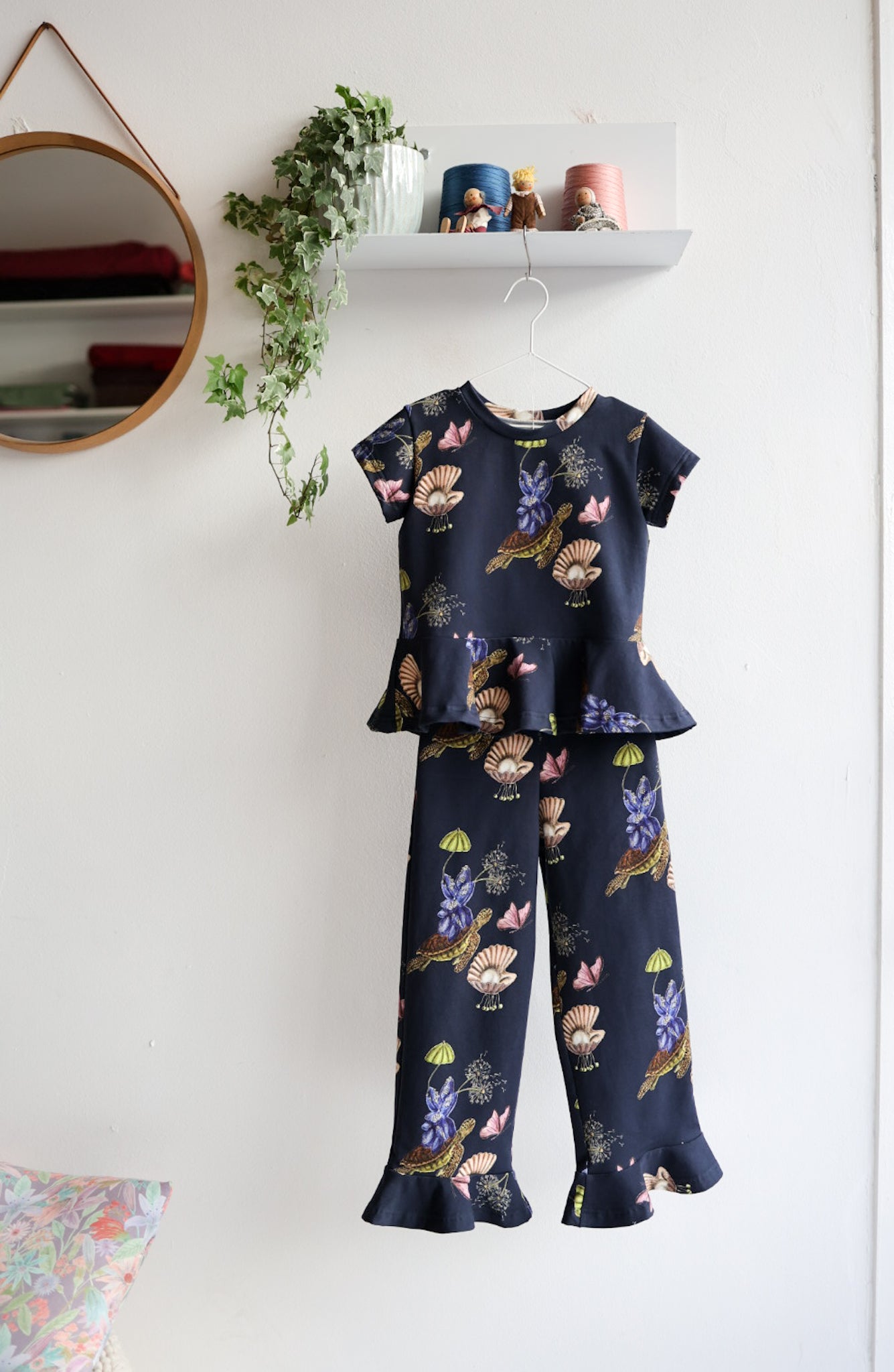 Navy blue nature-patterned kids pants and top hanging against a white wall with decorative items.