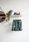 Teal kids skirt with nature-themed print hanging on a white wall.