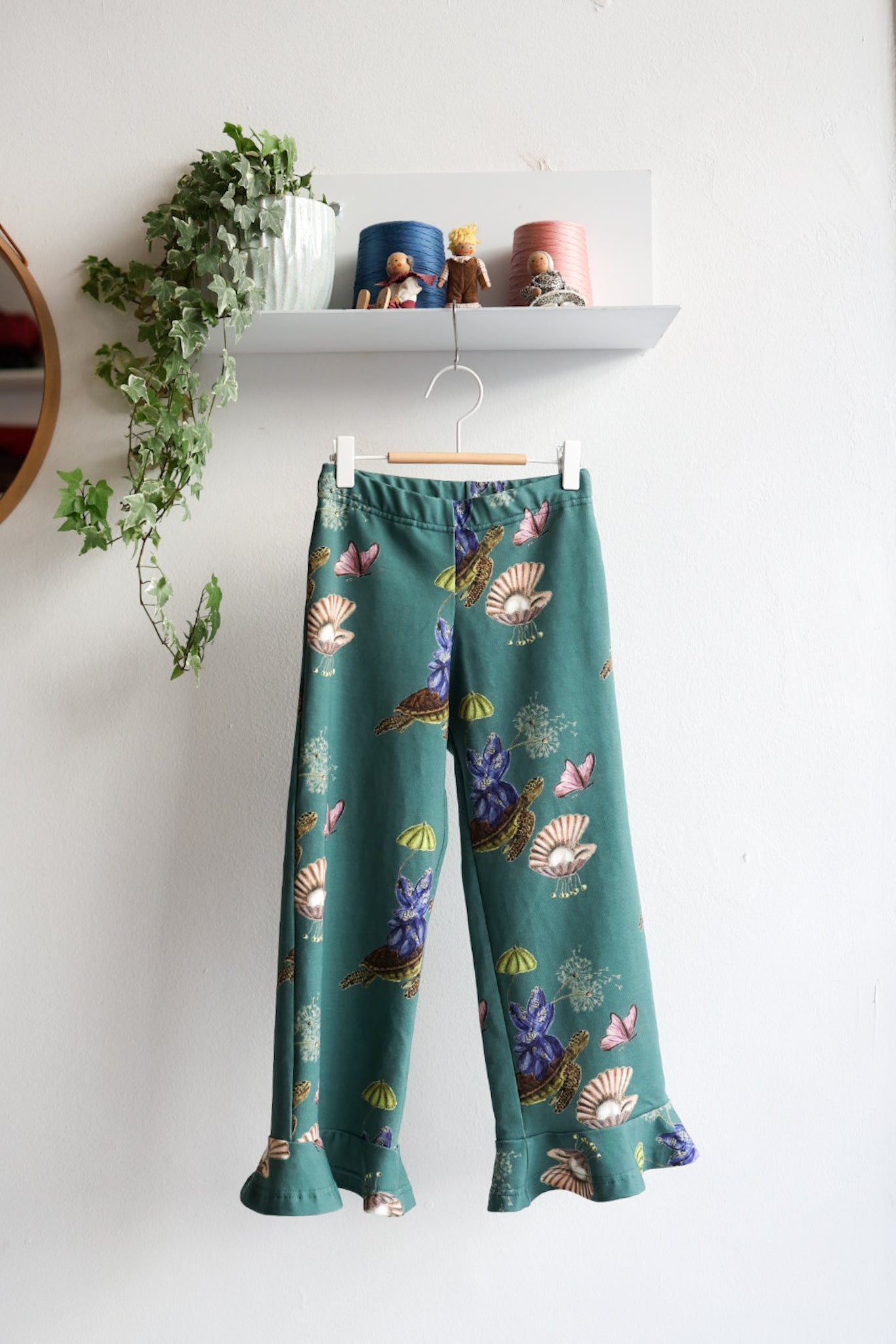 Teal nature-patterned kids pants hanging against a white wall with decorative items.