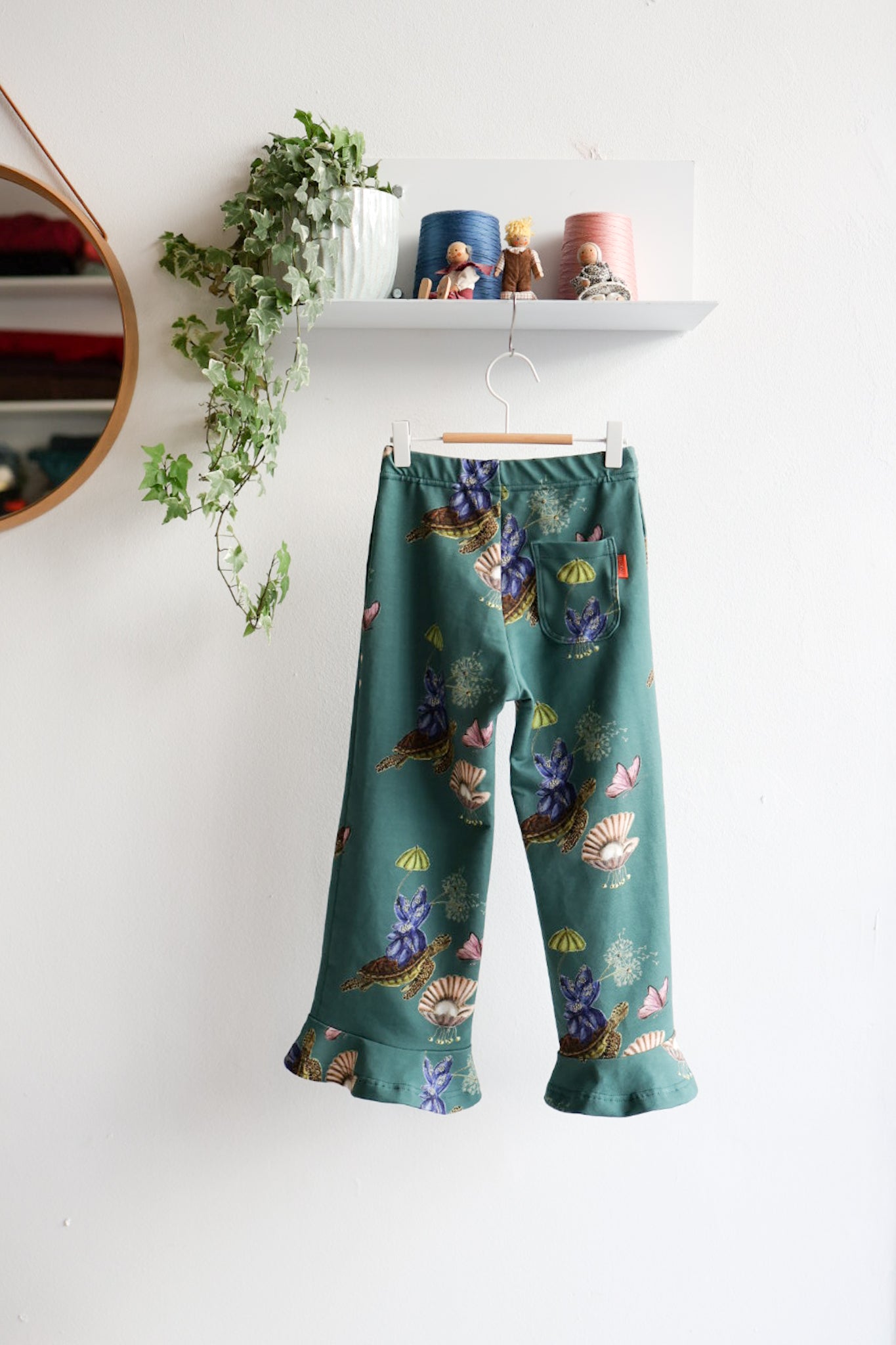 Teal nature-patterned kids pants hanging against a white wall with decorative items.