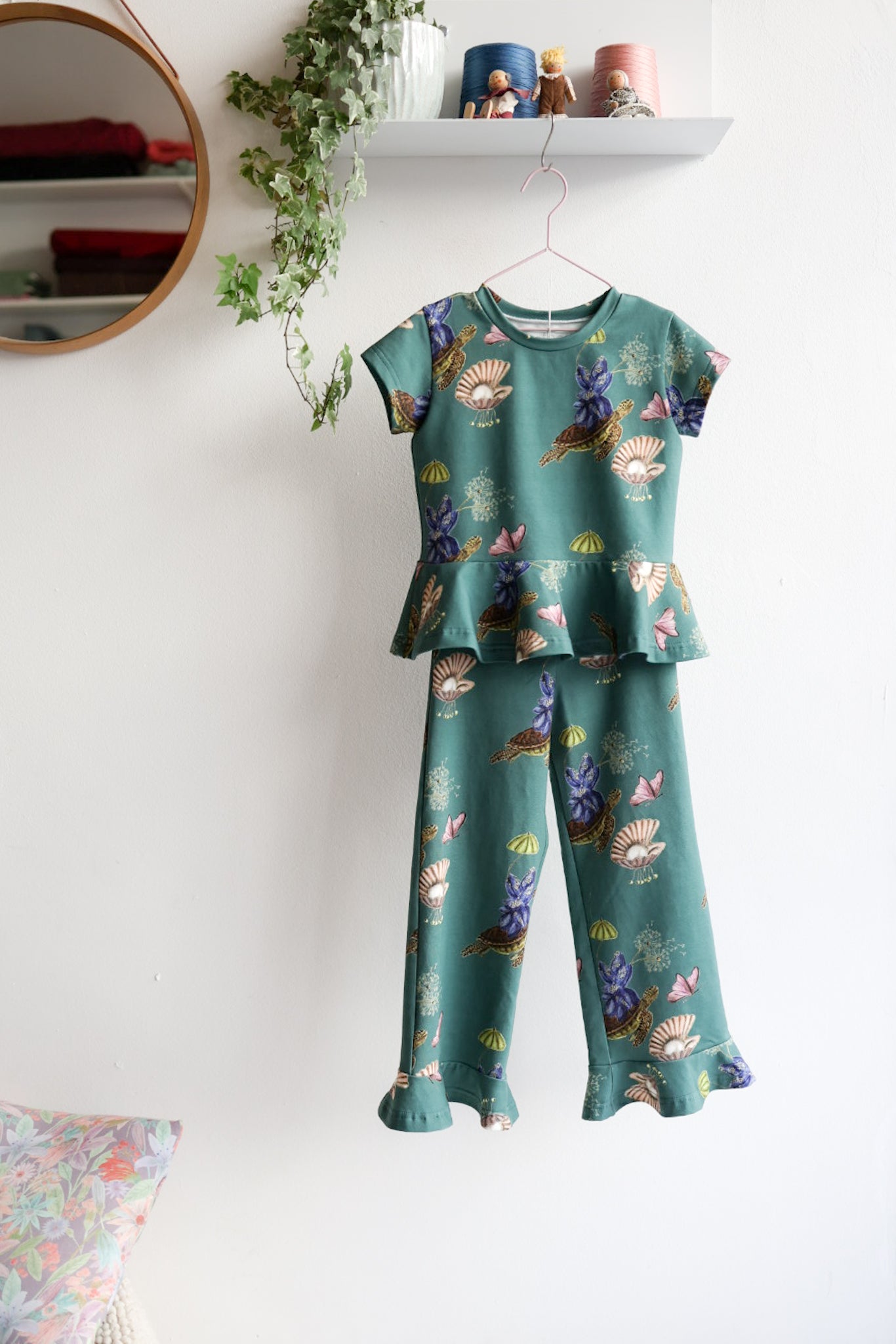 Teal nature-patterned kids pants and top hanging against a white wall with decorative items.