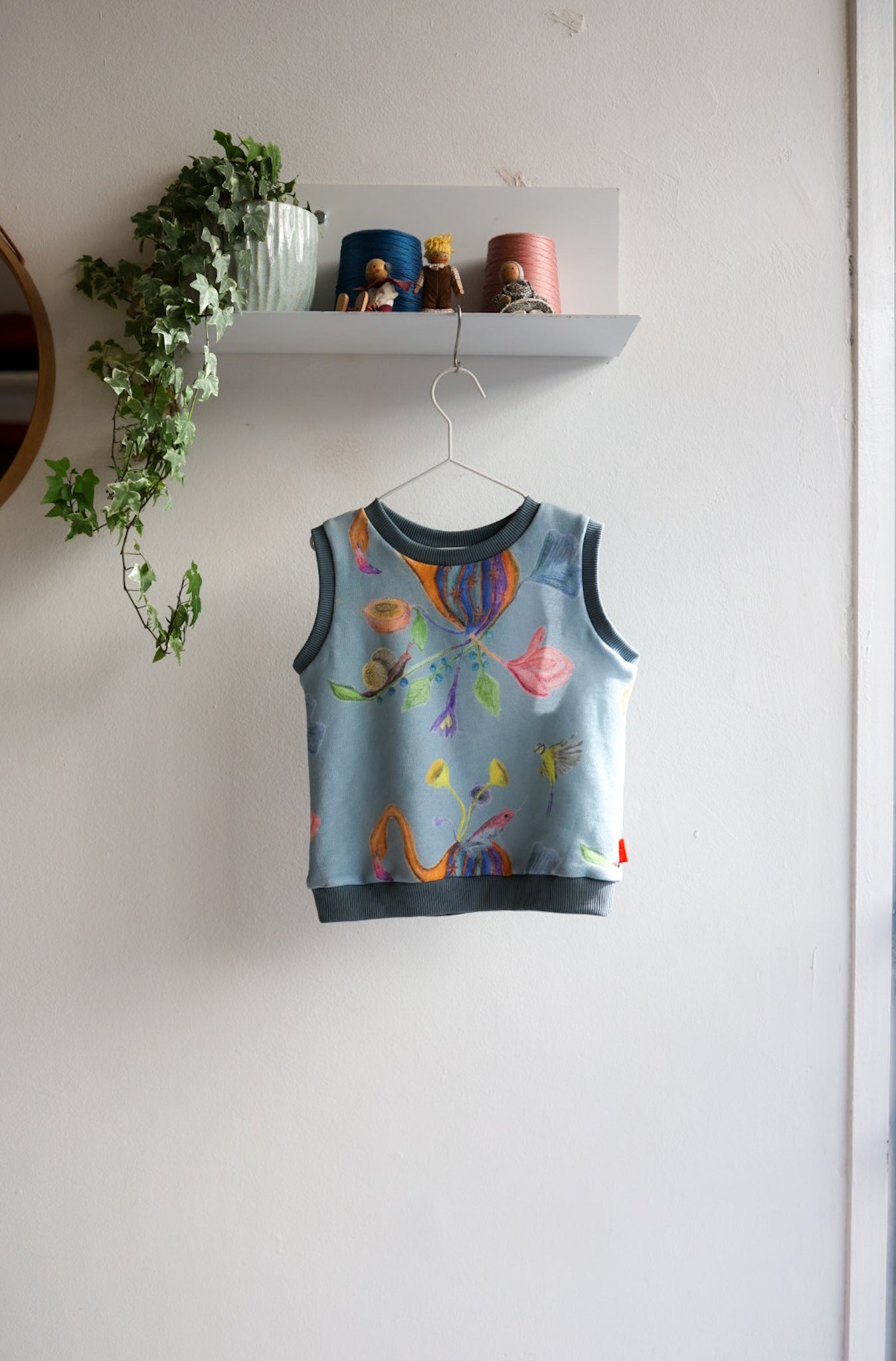 Colorful kids sleeveless top on a hanger against a white wall with a shelf and plant in the background.
