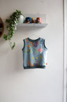 Colorful kids sleeveless top on a hanger against a white wall with a shelf and plant in the background.