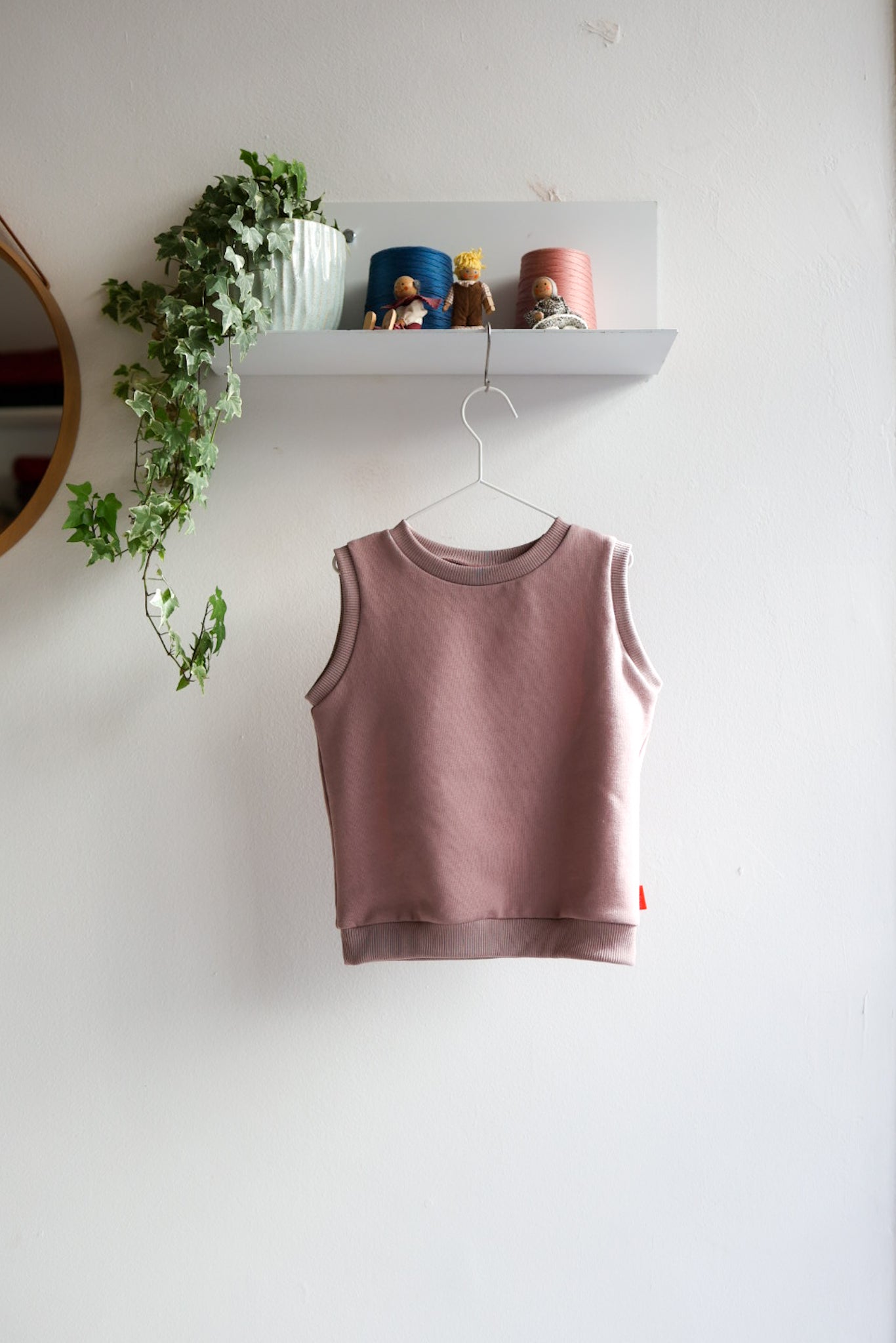 Pink kids sleeveless top hanging on a white wall with a shelf and plant in the background.