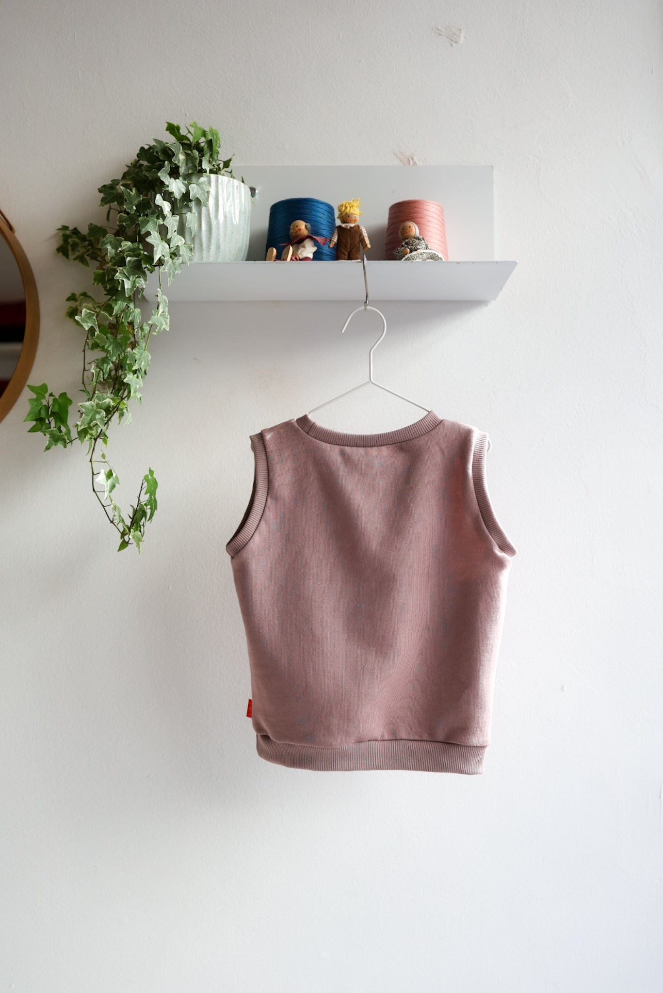 Pink kids sleeveless top on a hanger against a white wall with a shelf and plant in the background.