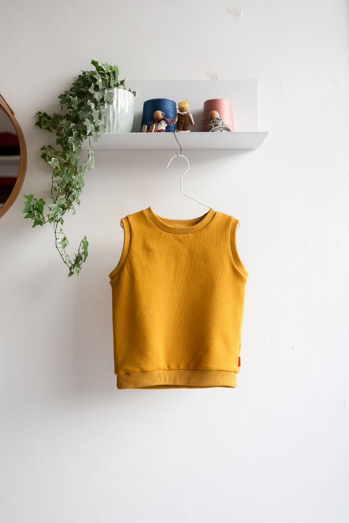Yellow kids sleeveless top hanging on a white wall with a shelf and plant in the background.