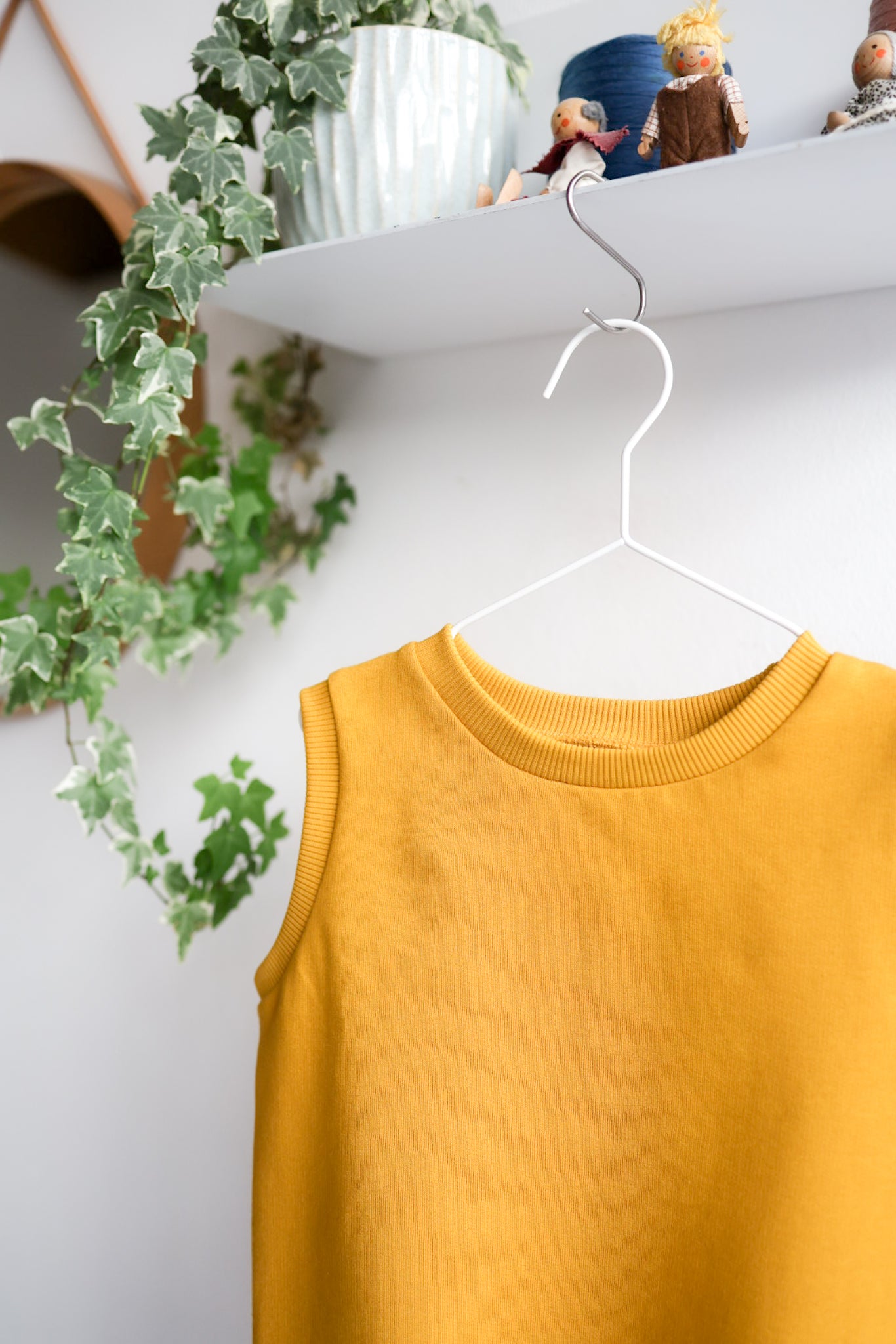 Mustard yellow kids sleeveless top on a hanger with a white shelf and plants in the background.