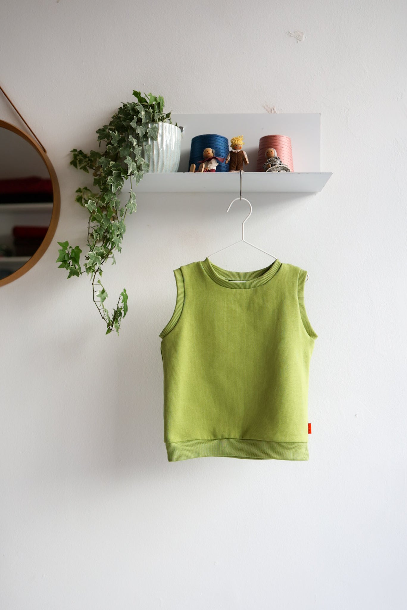 Green kids sleeveless top hanging on a white wall with a shelf and plant in the background.
