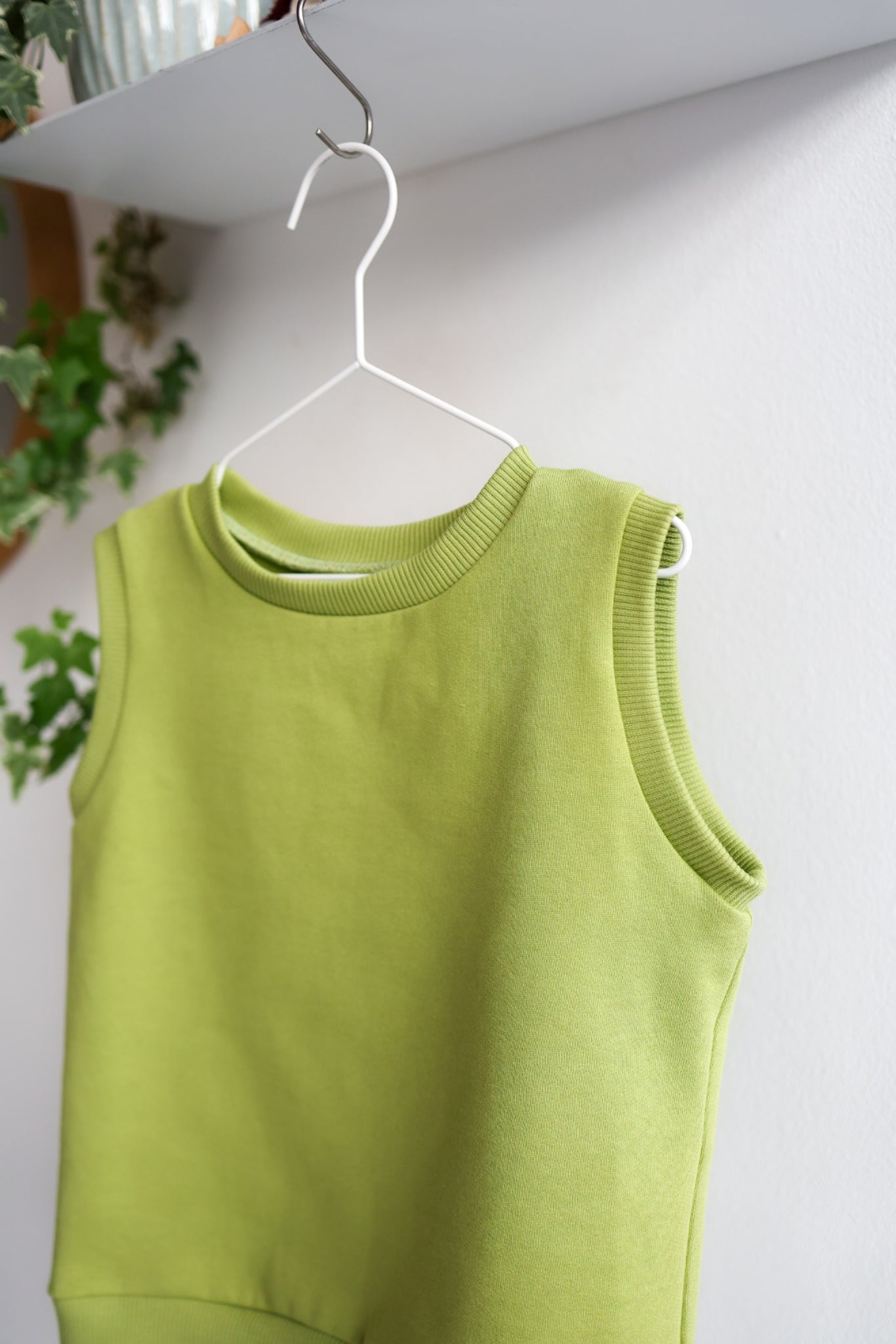 Green kids sleeveless top on a white hanger against a light gray wall.