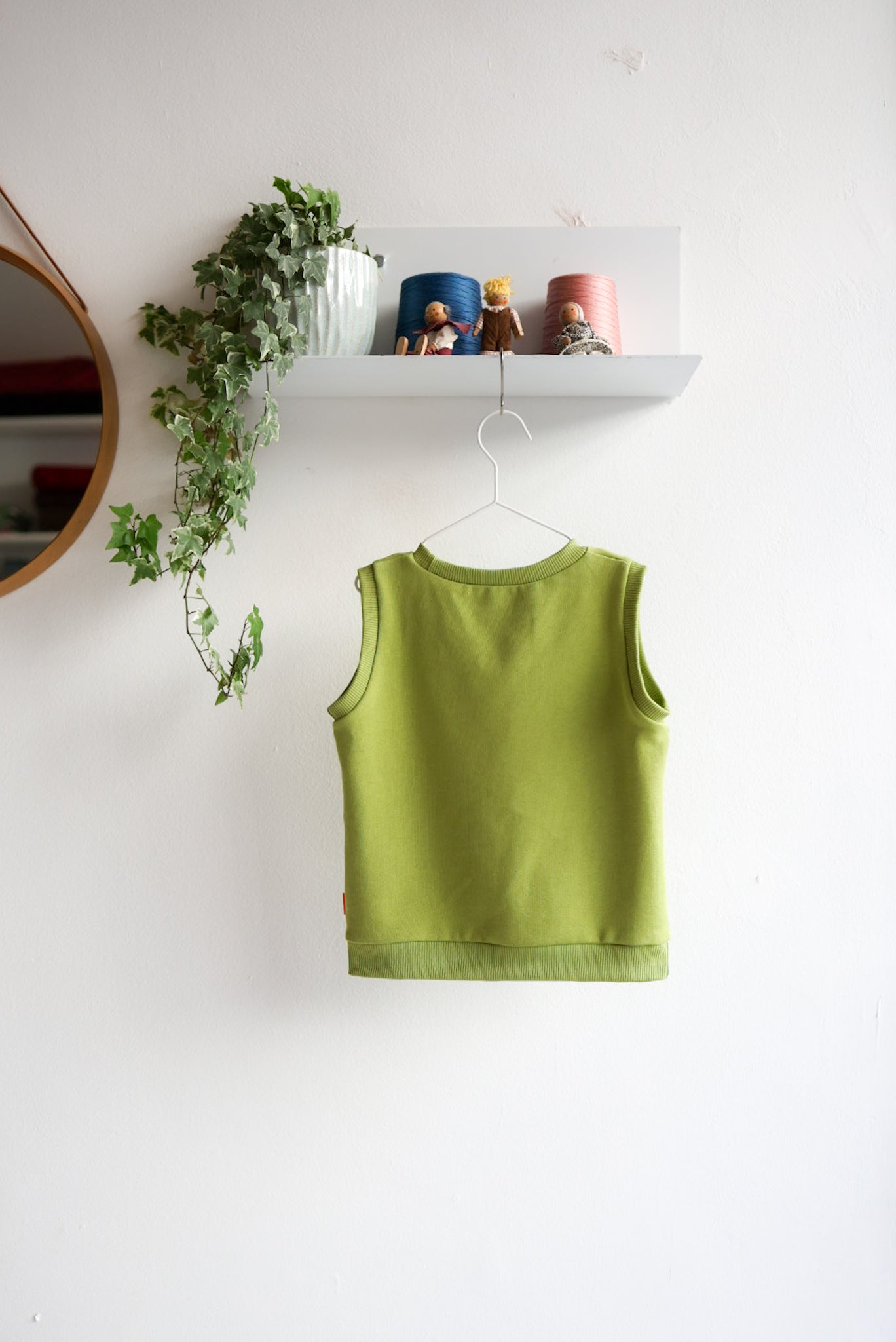 Green kids sleeveless top hanging on a white wall with a shelf and plants in the background.