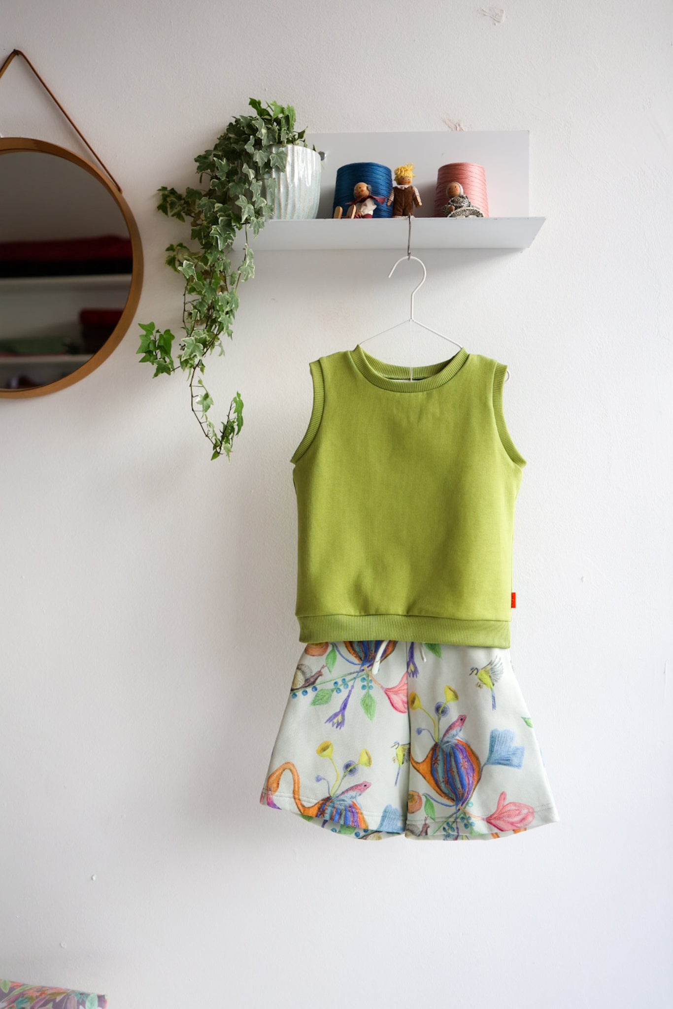 Green kids sleeveless top and colorful shorts on a white wall with a shelf and plant in the background.