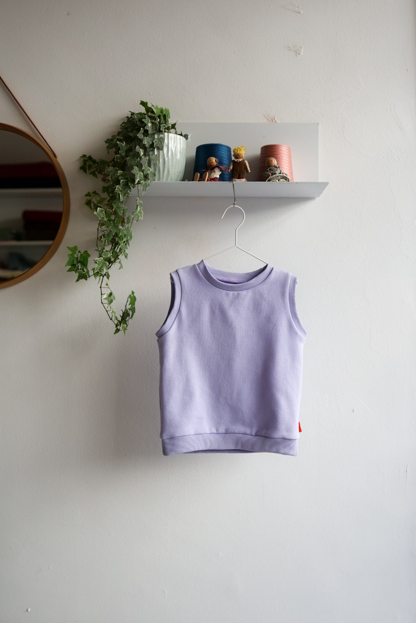 Purple kids sleeveless top hanging on a white wall with a shelf and decorative items in the background.