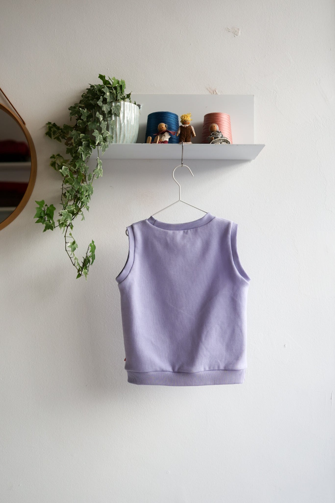 Purple kids sleeveless top on a hanger against a white wall with a shelf and plant in the background.