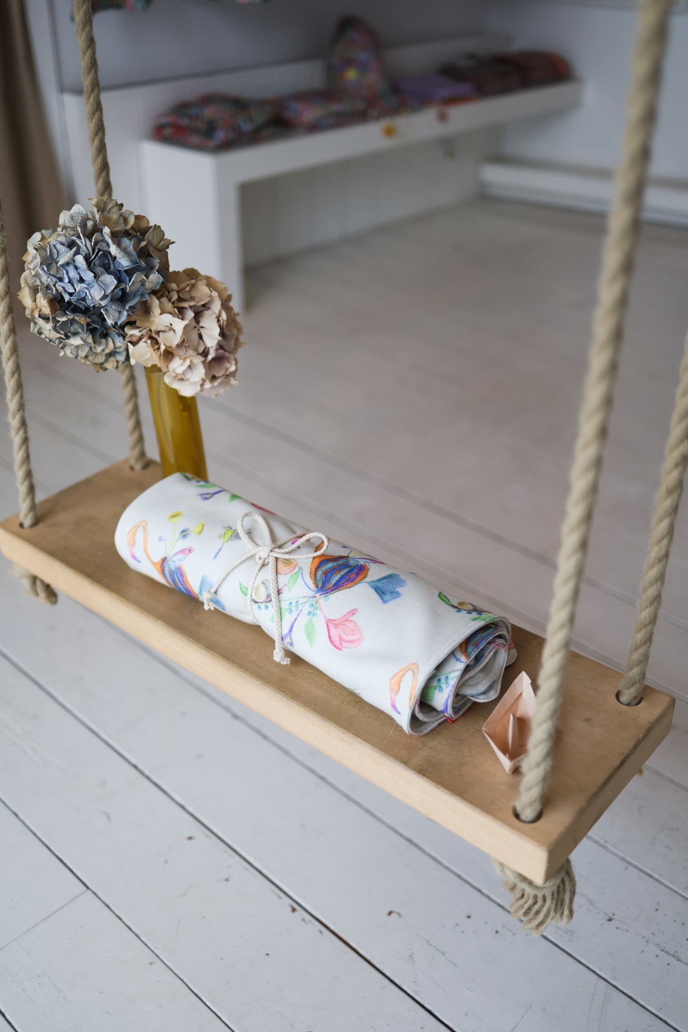 Wooden swing with a colorful babies blanket and decorative flowers on a white floor.