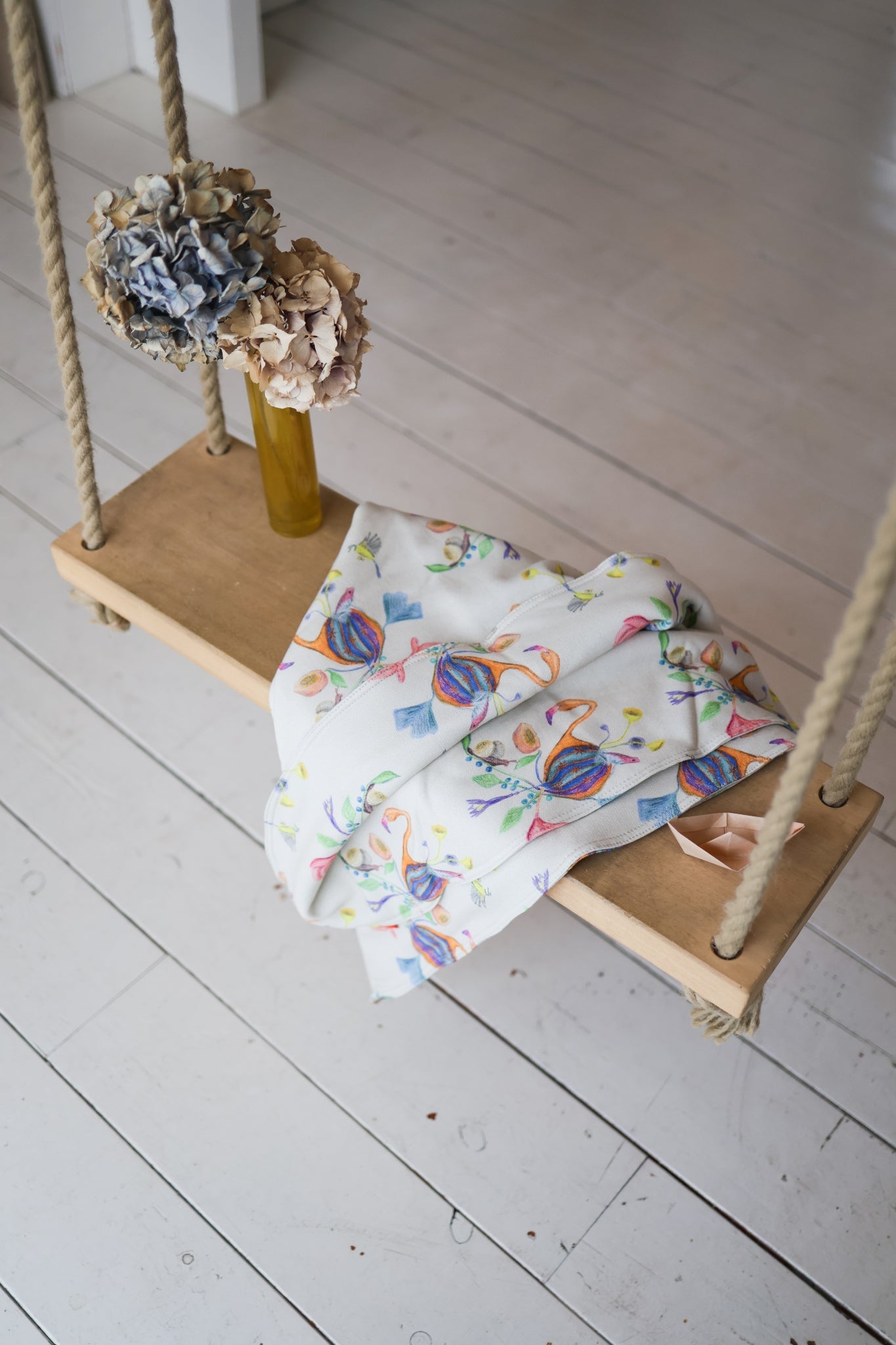 Wooden swing with a colorful babies blanket and decorative items on a light wooden floor.