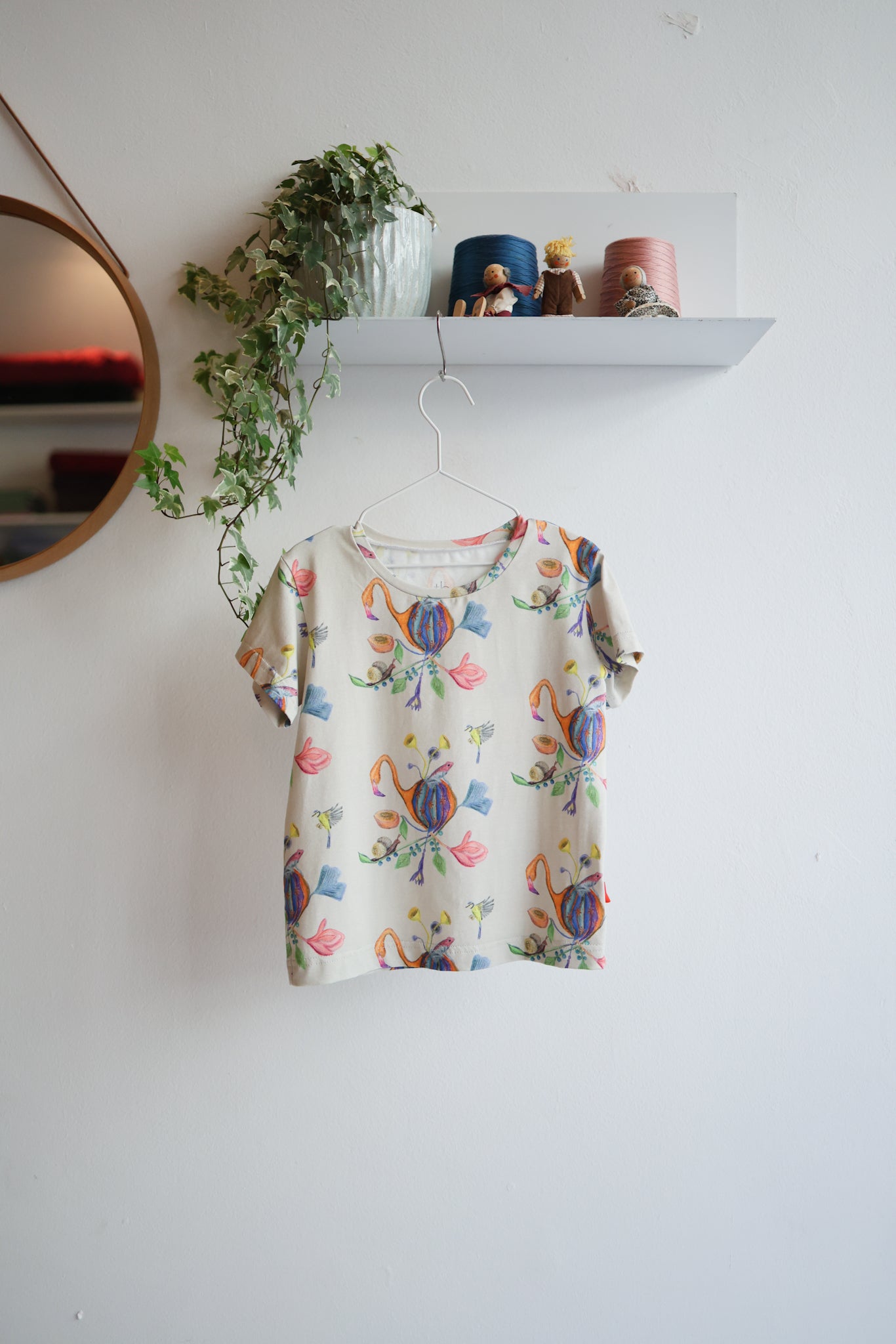 Colorful floral-patterned shirt on a hanger against a white wall with a shelf and plant in the background.