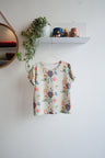 Colorful floral-patterned shirt on a hanger against a white wall with a shelf and plant in the background.