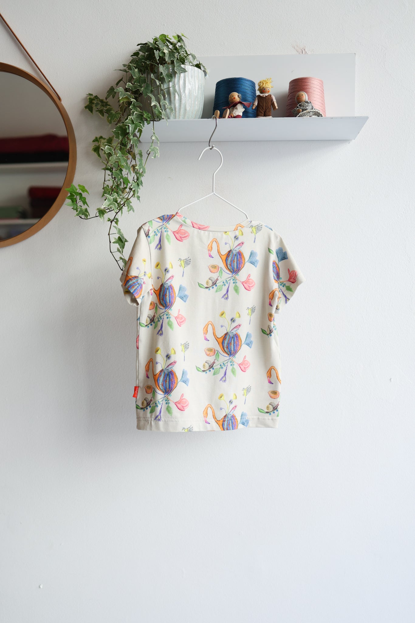 Colorful floral-patterned t-shirt hanging on a white wall with decorative items above.