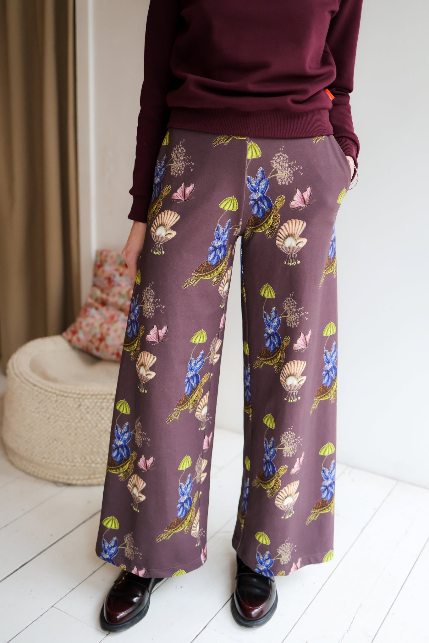 Woman wearing a sweater and patterned pants in a room with a white wall and wooden floor.