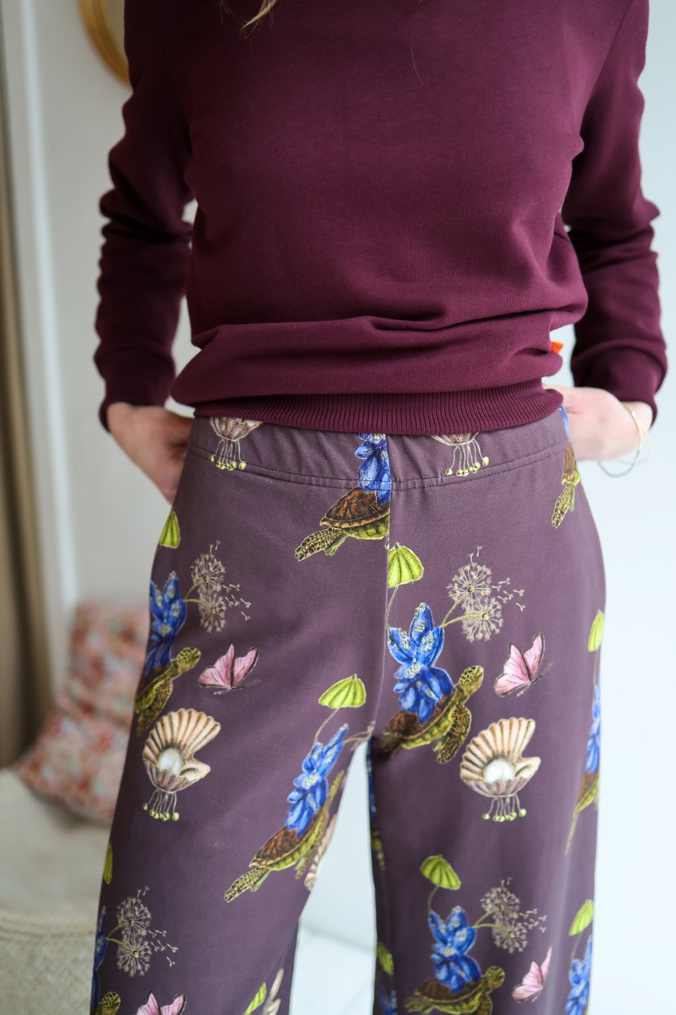 Woman wearing a sweater and patterned pants standing in a room with a blurred background.