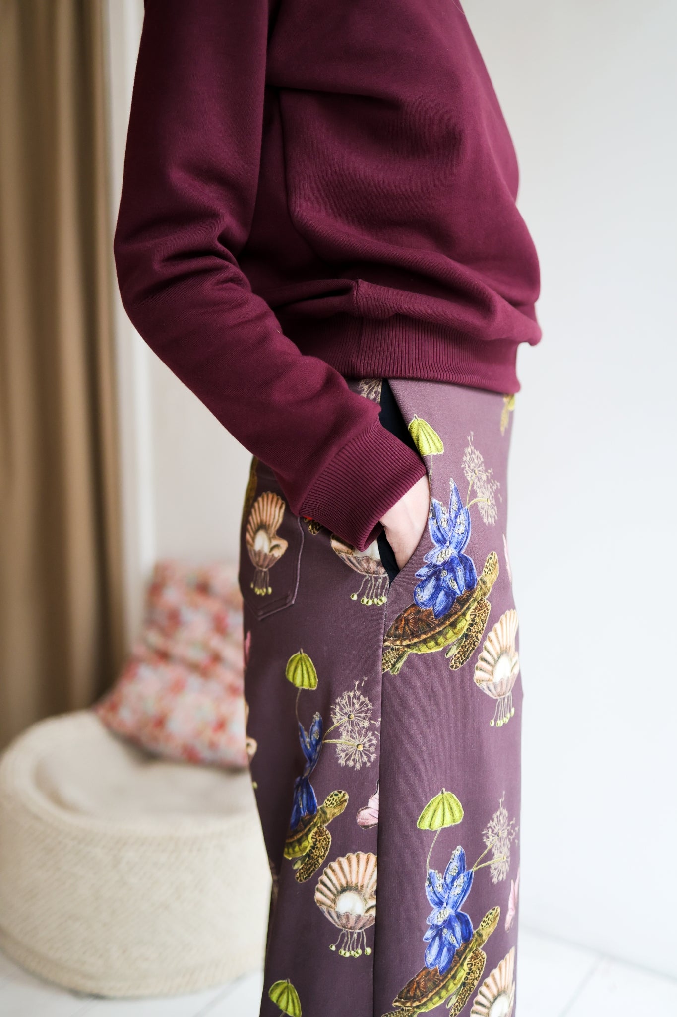 Woman wearing a sweater and patterned pants standing in a room with a blurred background.