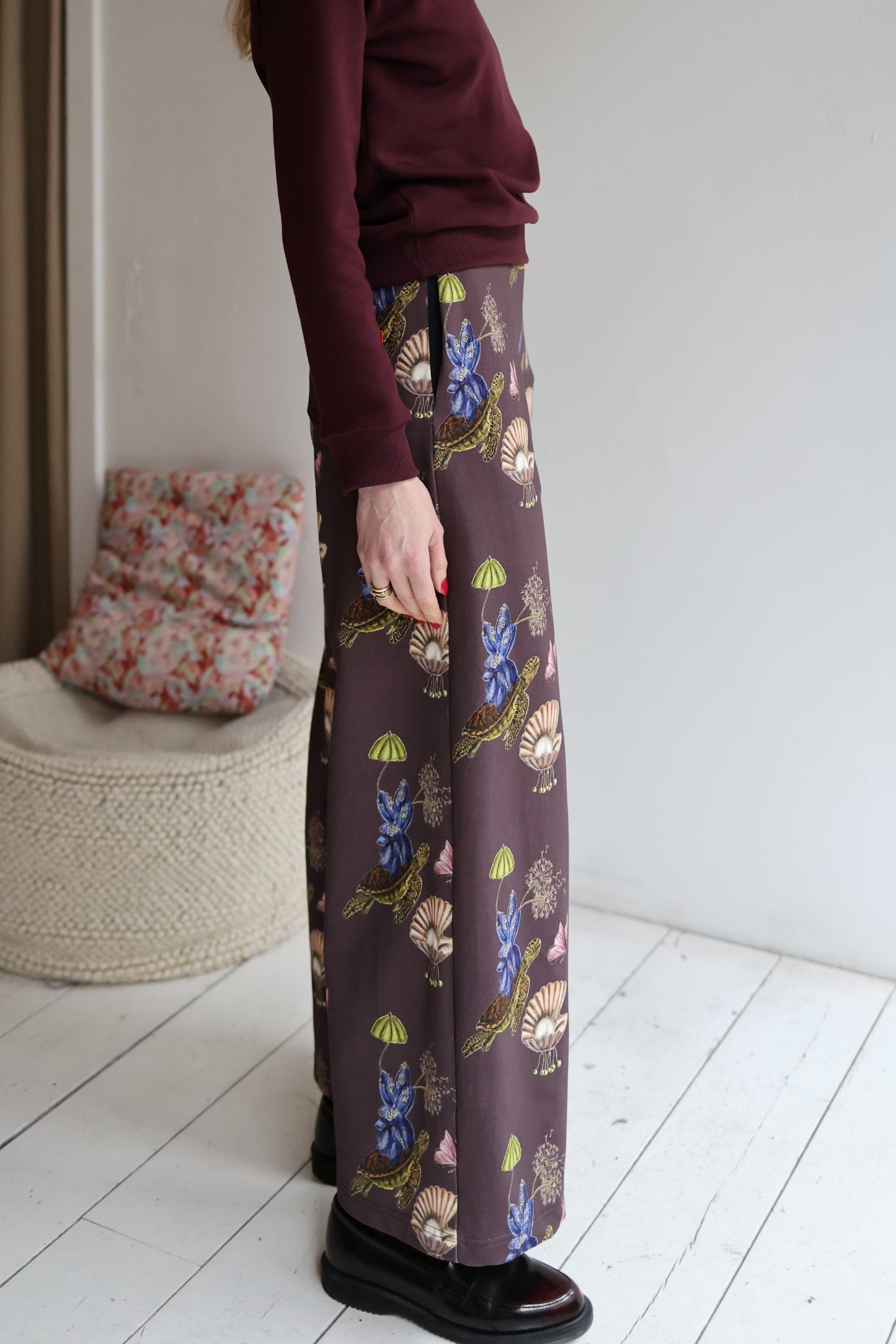 Woman wearing a sweater and patterned pants in a room with a white wall and wooden floor.