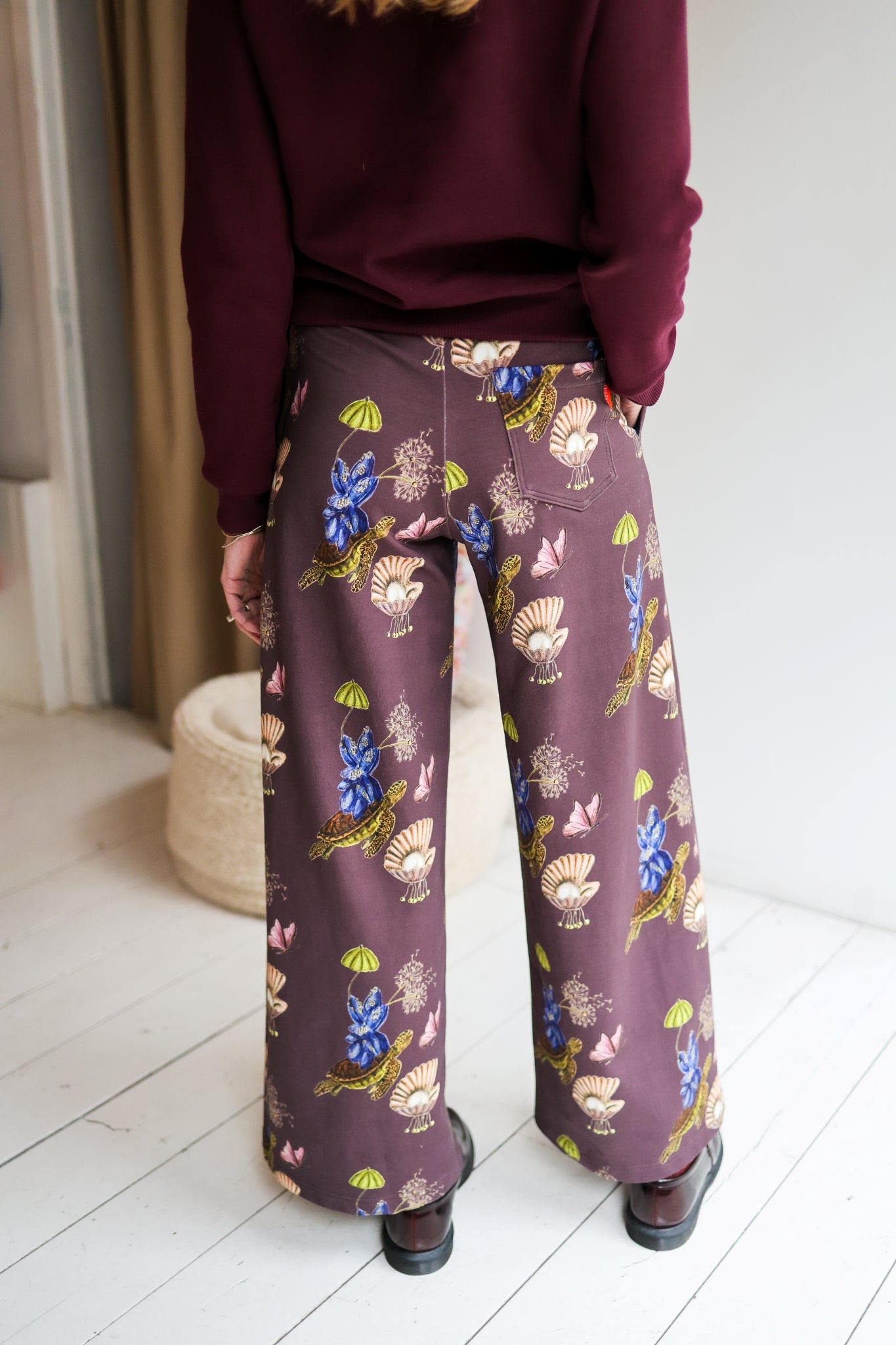 Woman wearing a sweater and patterned pants in a room with a white wall and wooden floor.