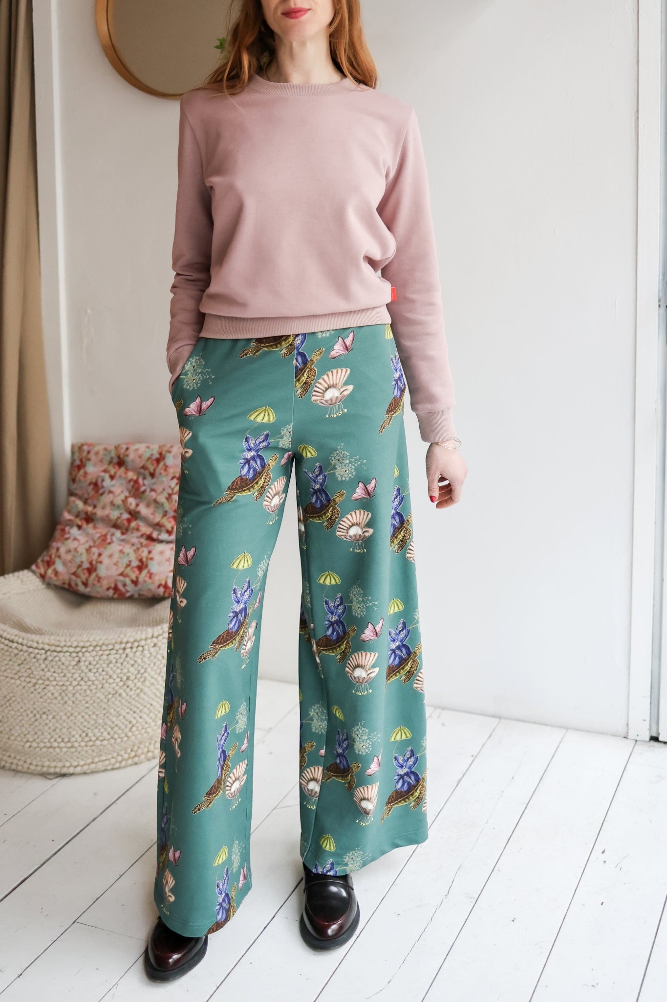 Woman wearing a pink sweater and teal floral pants in a room with a white wall and wooden floor.