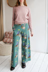 Woman wearing a pink sweater and teal floral pants in a room with a white wall and wooden floor.