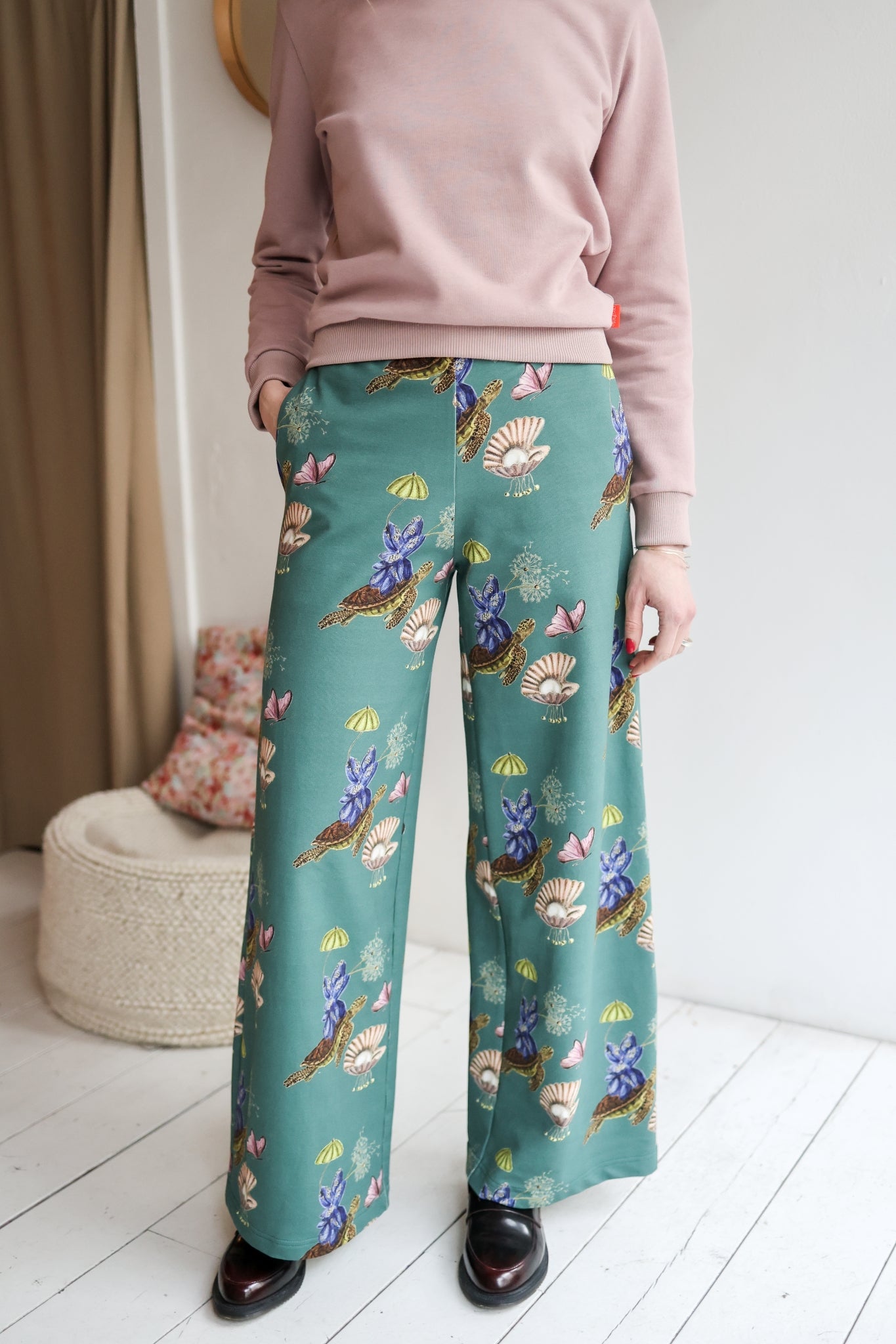 Woman wearing a pink sweater and teal floral pants in a room with a white wall and wooden floor.